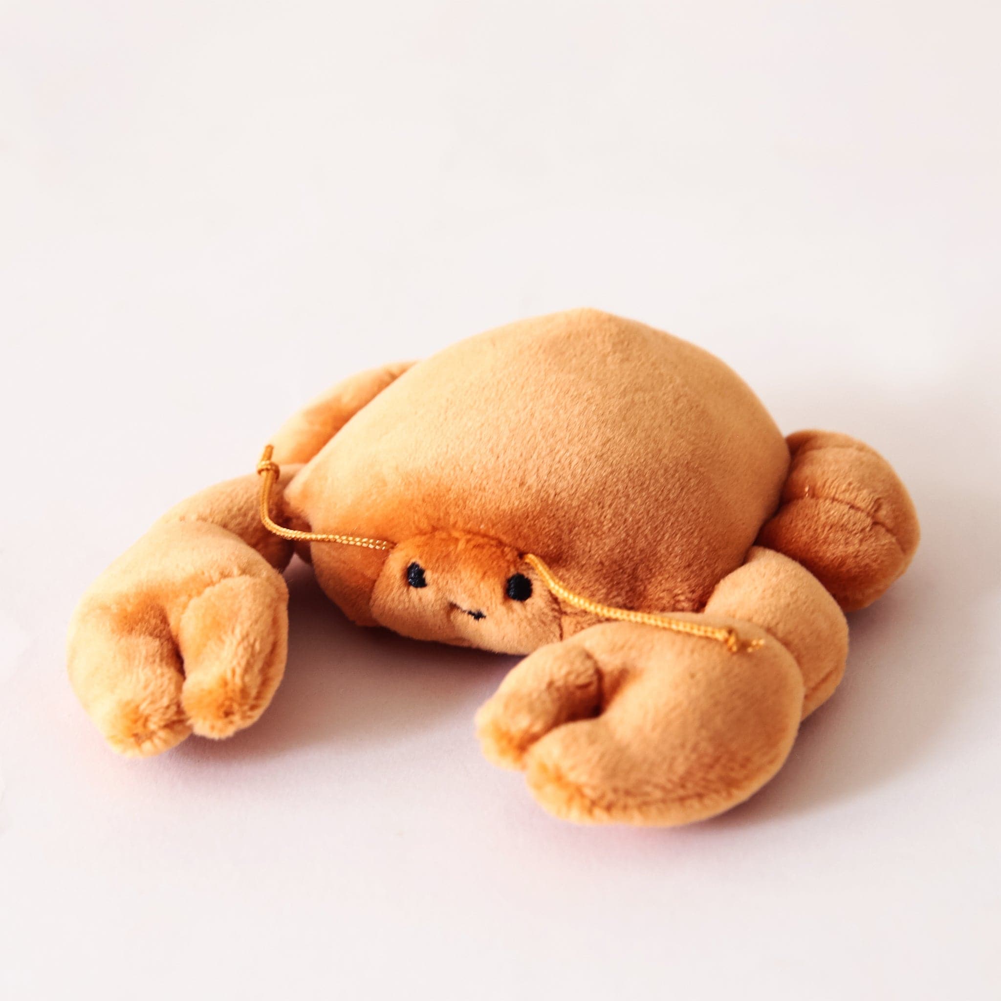 In front of a cream background is a stuffed orange crab laying on its stomach. It has two large claws in the front and two little claws in the back. On its face are two black eyes and a little black smile. He has two orange antennas above his eyes.