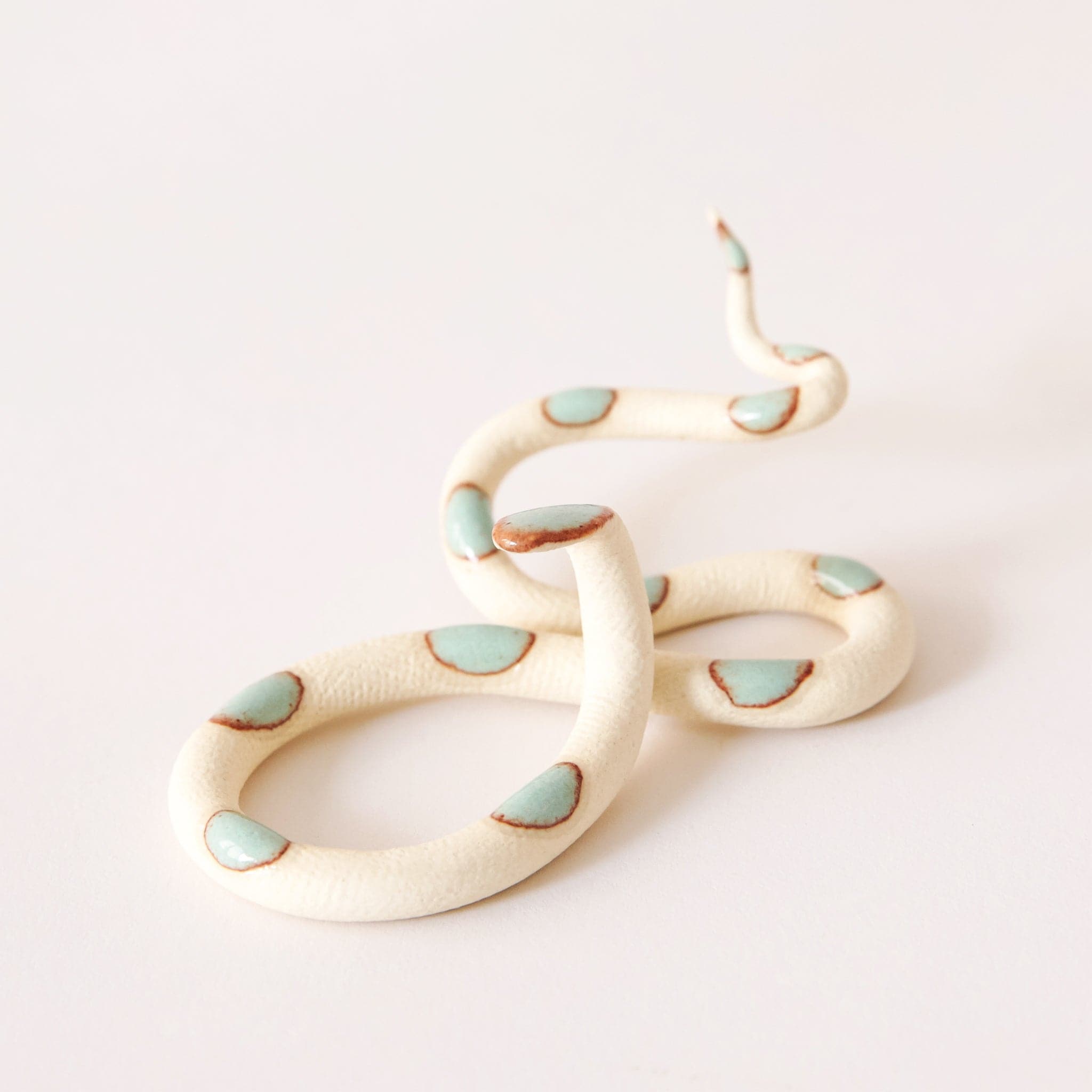 Sitting against a white background is a ceramic, cream snake. The snake&#39;s body twists multiple times from head to tail. There are brown bordered turquoise circles on the top of the snake. 