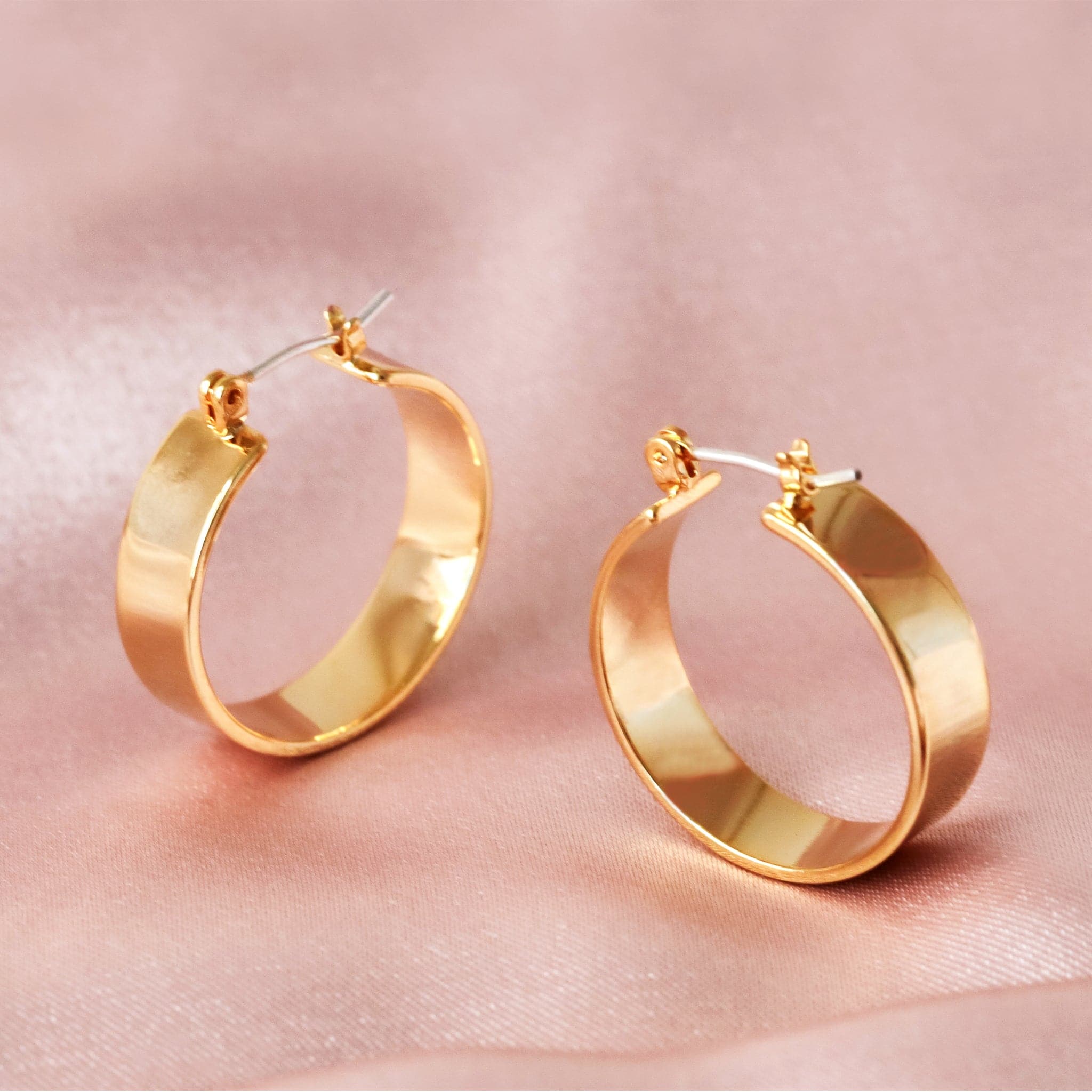 Medium sized gold hoops that have a wider frame and a thin depth. They feature a straight post and a clasp on the back top of the hoop to secure it on your ear.