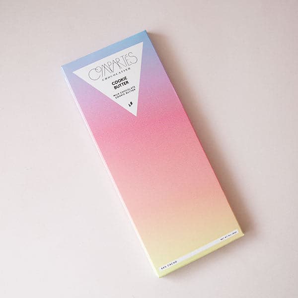 A rectangle bar of chocolate with a gradient wrapper that goes from blue to purple, to pink, to orange, to yellow along with a upside down triangle shape and text in the center that reads, "Compartes Cookie Butter Milk Chocolate".