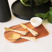 An arched wood cutting board, photographed with a bowl and serving utensils that have a wicker handle and gold metal piece (not included with purchase).