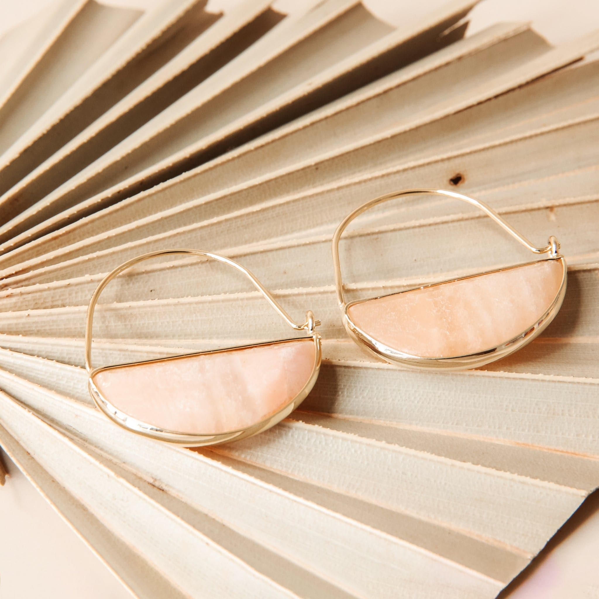 Laying on top of a dried palm frond is a pair of gold earrings. The top is a thin gold hoop. The bottom is a peach stone half circle with a gold border. 