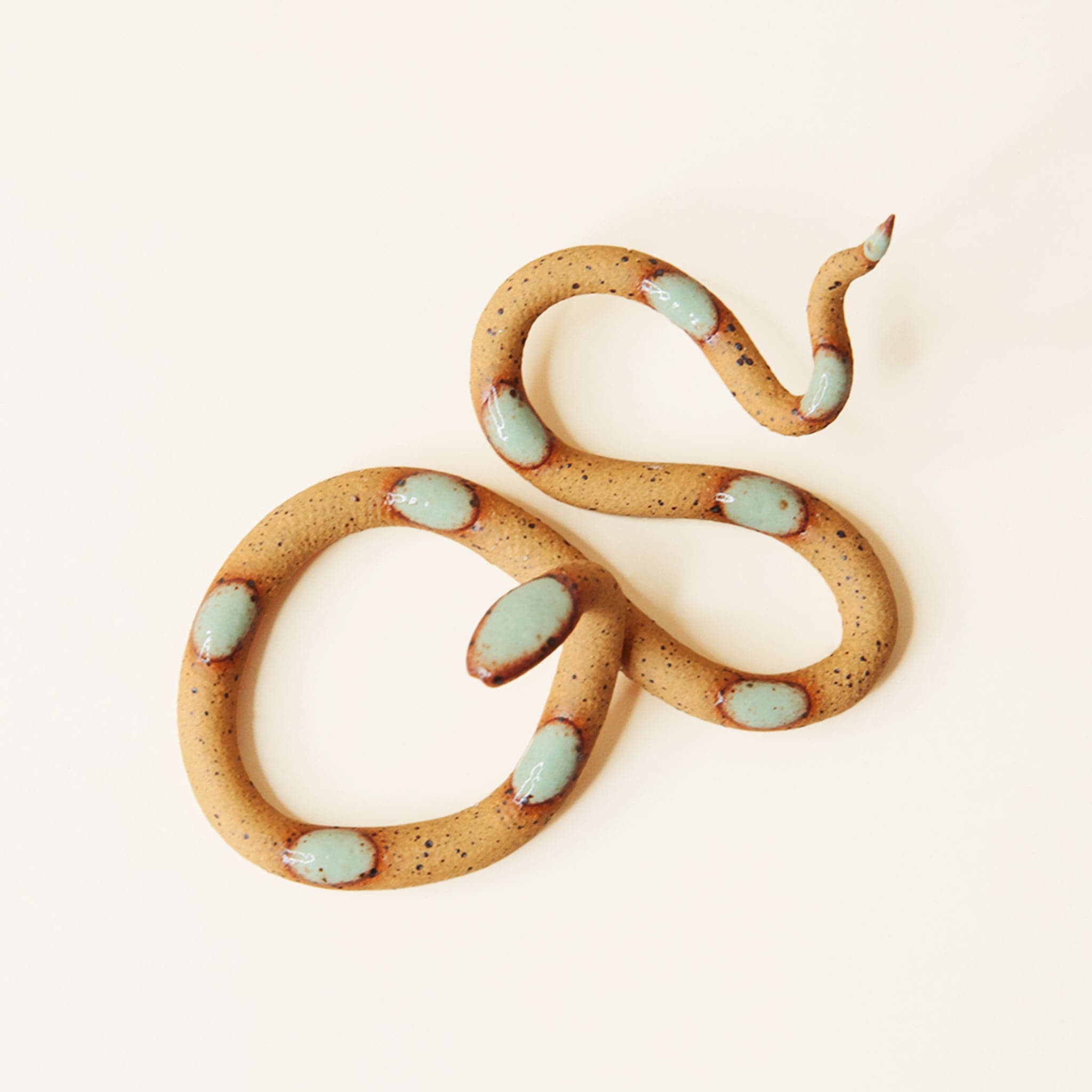 In front of a tan background is a brown ceramic snake. The snake is speckled light brown with light blue spots. 