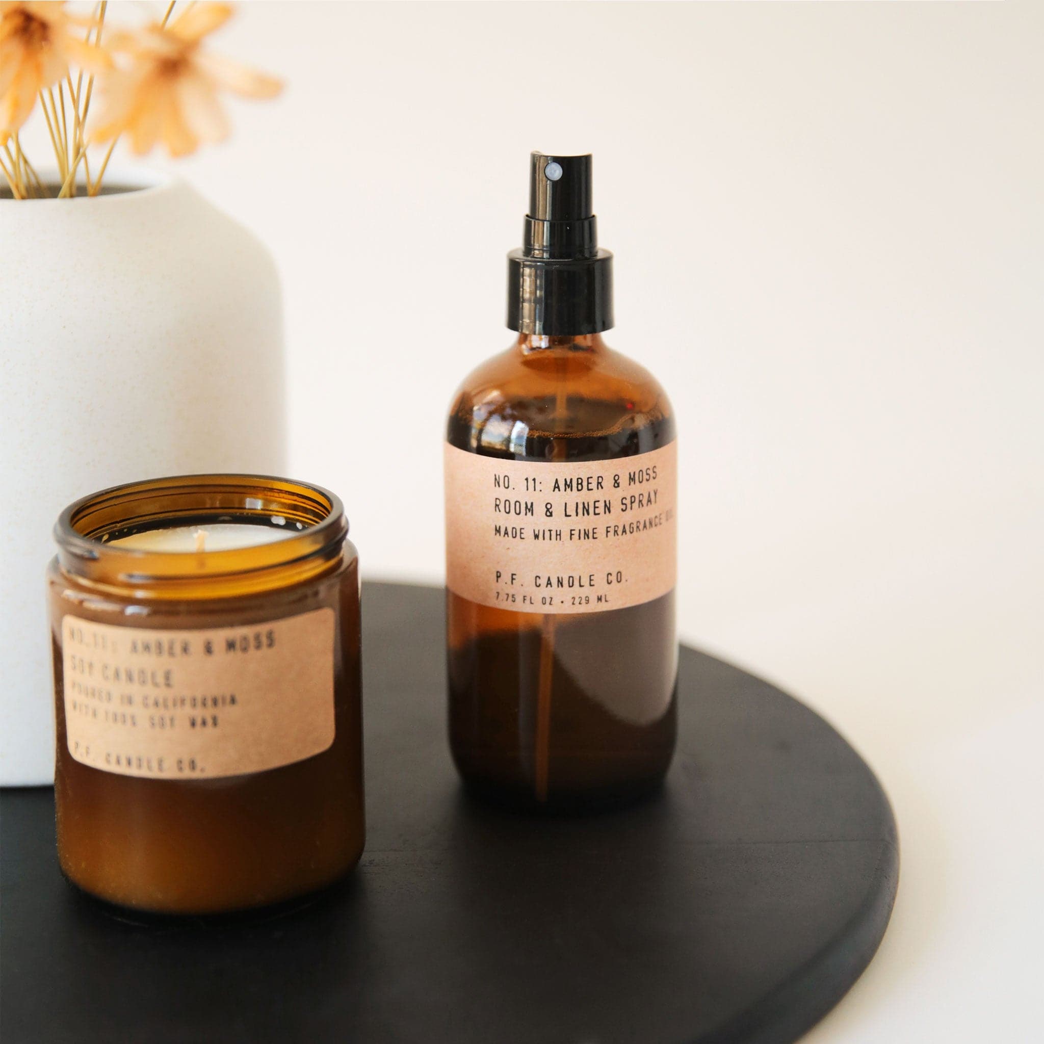 An amber glass spray bottle with a neutral brown label that reads, "No. 11: Amber & Moss Room & Linen Spray. P.F. Candle Co." staged on a black tray with a candle to the left of it.