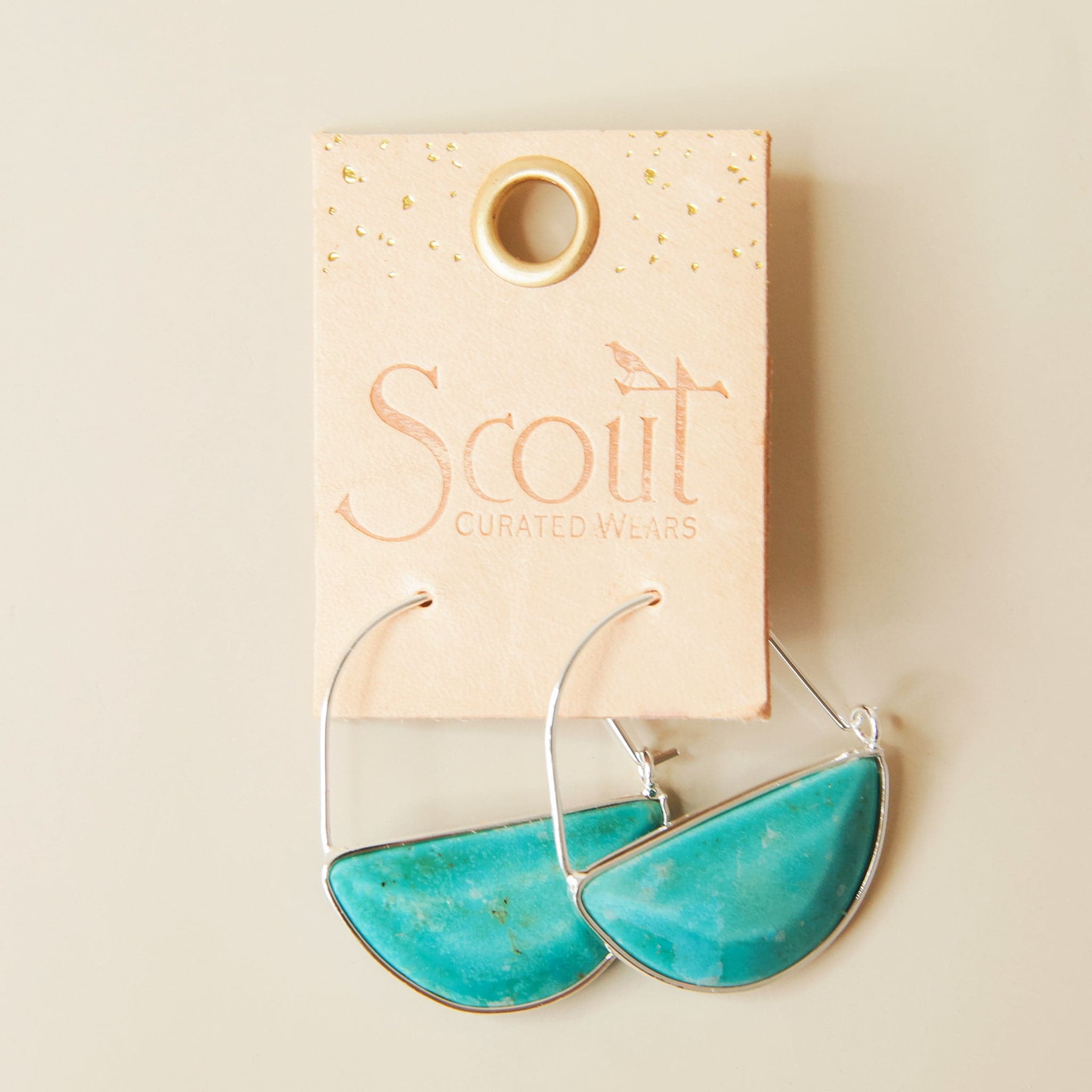 In front of a cream background is a pair of silver hoops. The bottom half of the hoop is a turquoise stone. The hoops are on a peach leather tag. Etched into the tag is text that reads ‘Scout curated wears.'