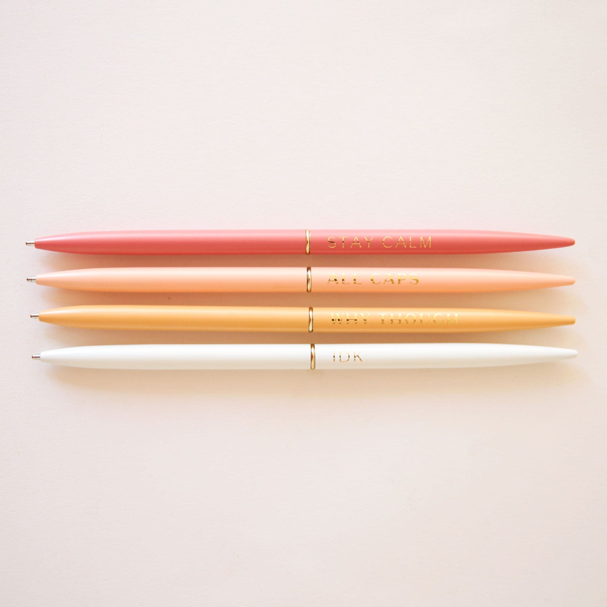 A set of 4 slender pens in yellow, white, salmon and rust. Each pen says something different. The rust pen says, "Stay Calm". The salmon pen says, "All Caps". The white pens says, "Idk". And the yellow pen says, "Why Though".