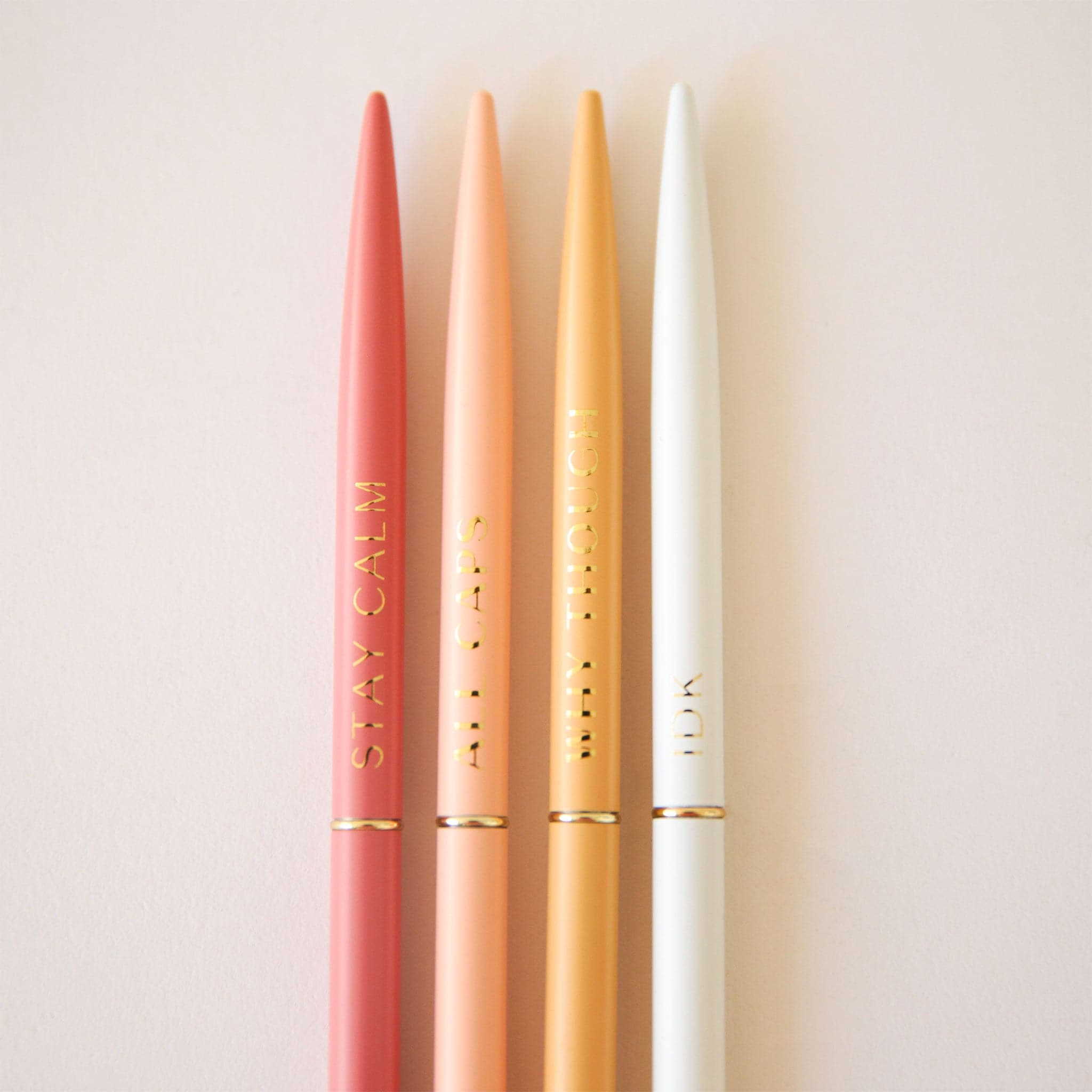 A set of 4 slender pens in yellow, white, salmon and rust. Each pen says something different. The rust pen says, "Stay Calm". The salmon pen says, "All Caps". The white pens says, "Idk". And the yellow pen says, "Why Though".