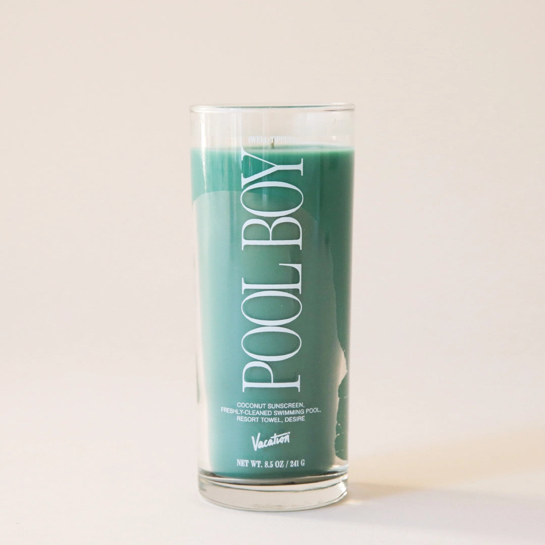 A thin clear glass candle with a green wax candle inside and has white text that reads, "Pool Boy". 