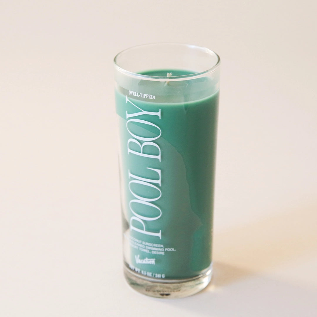 A thin clear glass candle with a green wax candle inside and has white text that reads, "Pool Boy".