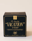 A black glass candle jar with a white soy wax blend on the inside. There is a gold label on the front of the candle that says, "The Legendary "Vacation" by Vacation".
