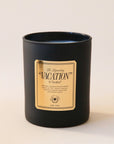 A black glass candle jar with a white soy wax blend on the inside. There is a gold label on the front of the candle that says, "The Legendary "Vacation" by Vacation".