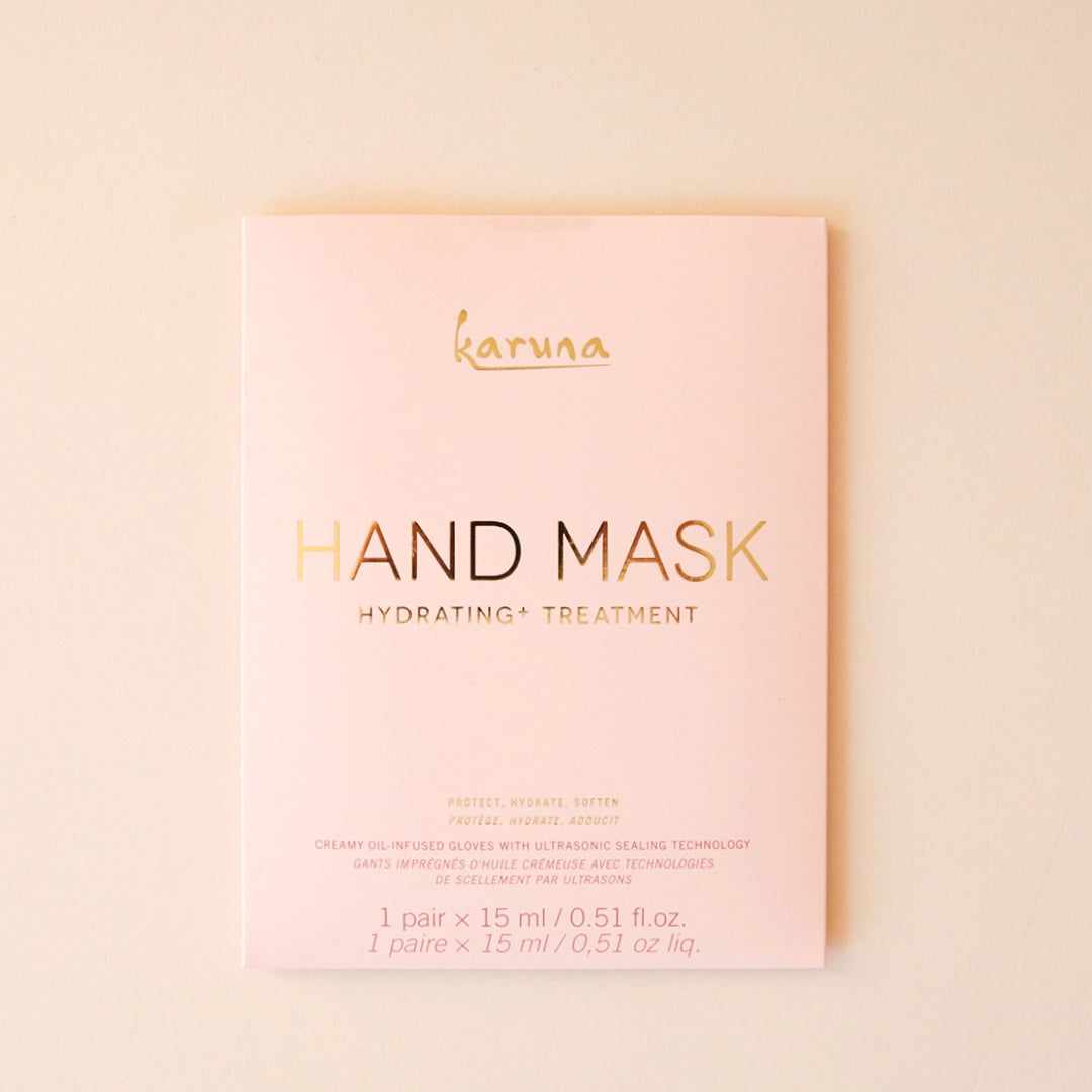 On a cream background is a pink packet with a hand mask inside that reads, &quot;Karuna Hand Mask Hydrating + Treatment&quot;.