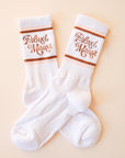White crew socks with "Proud Mama" written in brown swirly text along with two brown and light pink stripes above and below it.