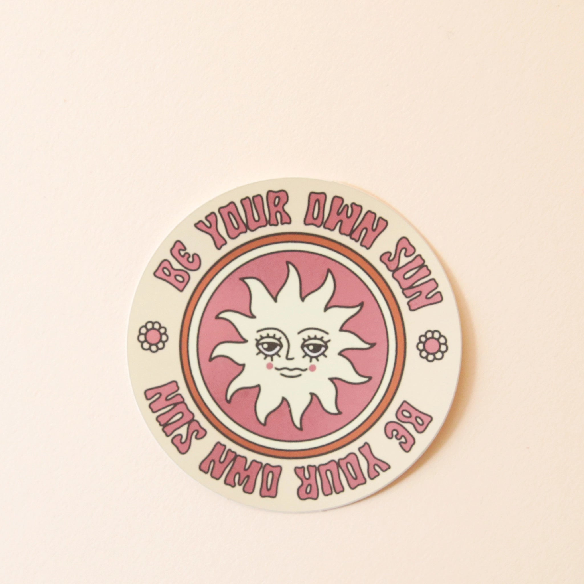 On a cream background is a cream and pink circle sticker with a sun illustration in the center and text around the edge of the circle that reads, &quot;Be Your Own Sun&quot;. 