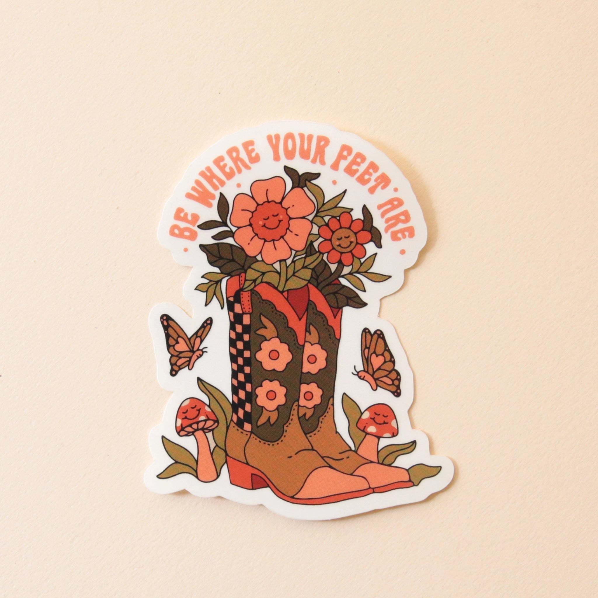 On a white background is a sticker shaped in a cowgirl boot with flowers and surrounded by mushrooms and butterflies as well as text arched above that reads, "Be Where Your Feet Are".
