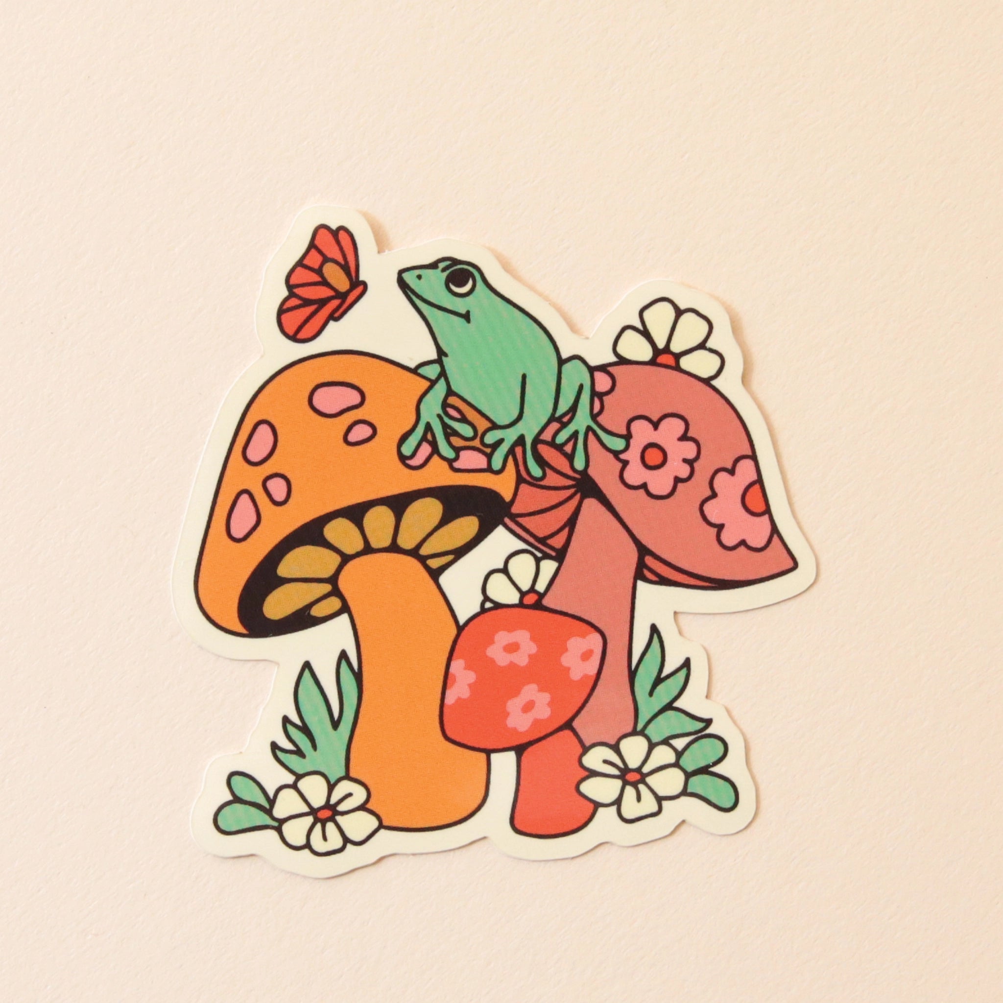 On a cream background is a sticker with an illustration of multi colored mushrooms with a green frog siting on top of it. 