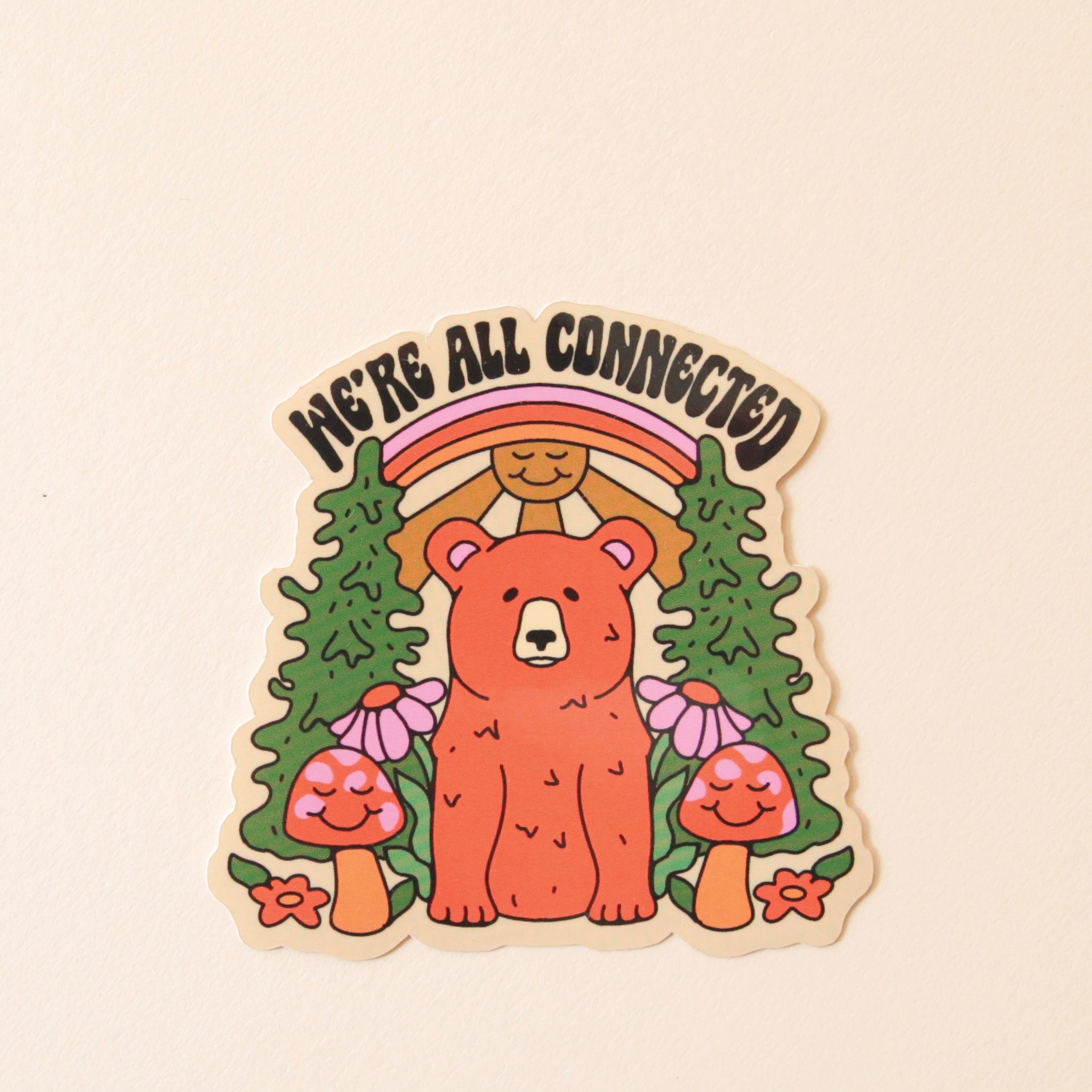 On a cream background is a sticker with a bear surrounded by trees, mushrooms and flowers while underneath an illustration of a smiling sun and rainbow and black text at the top that reads, &quot;We&#39;re All Connected&quot;.