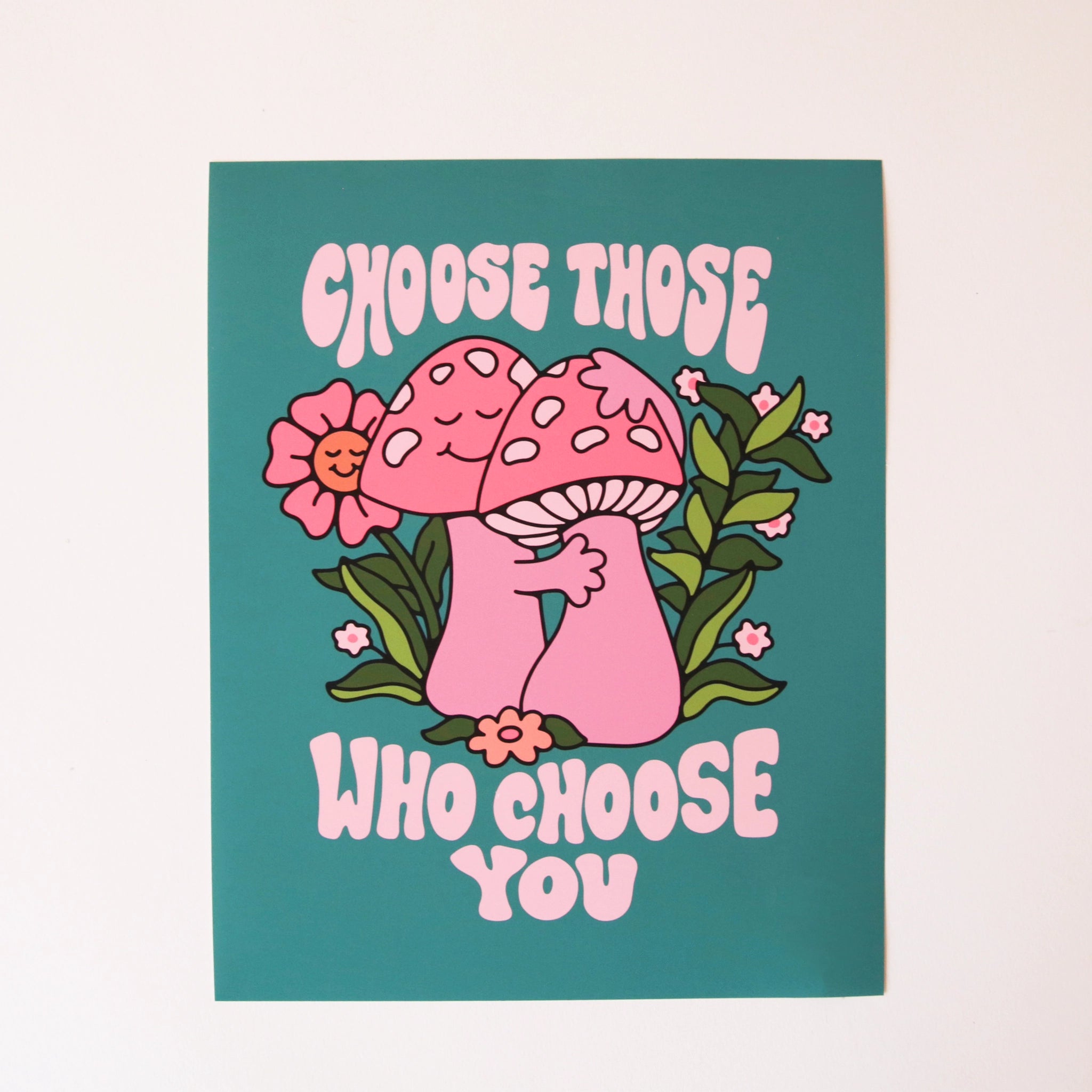 A turquoise blue art print featuring two pink mushrooms giving each other a hug along with a pink smiling daisy in the background, green vines and text above and below the graphic that reads, "Choose Those Who Choose You" in light pink.
