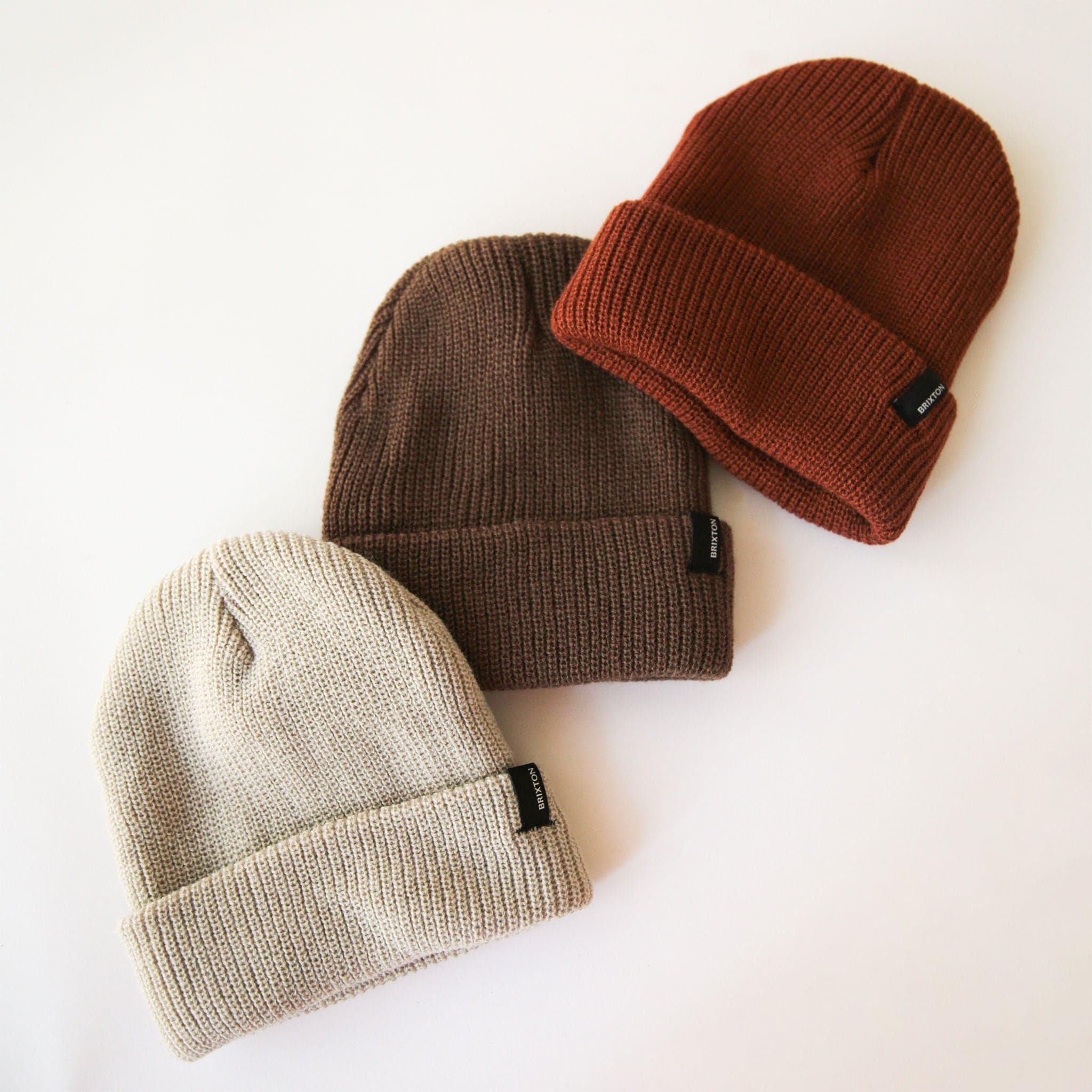 On a cream background is a brown ribbed beanie with a small black Brixton label on the edge of the folded brim photographed with a rust colored beanie and a neutral beige colored beanie. 
