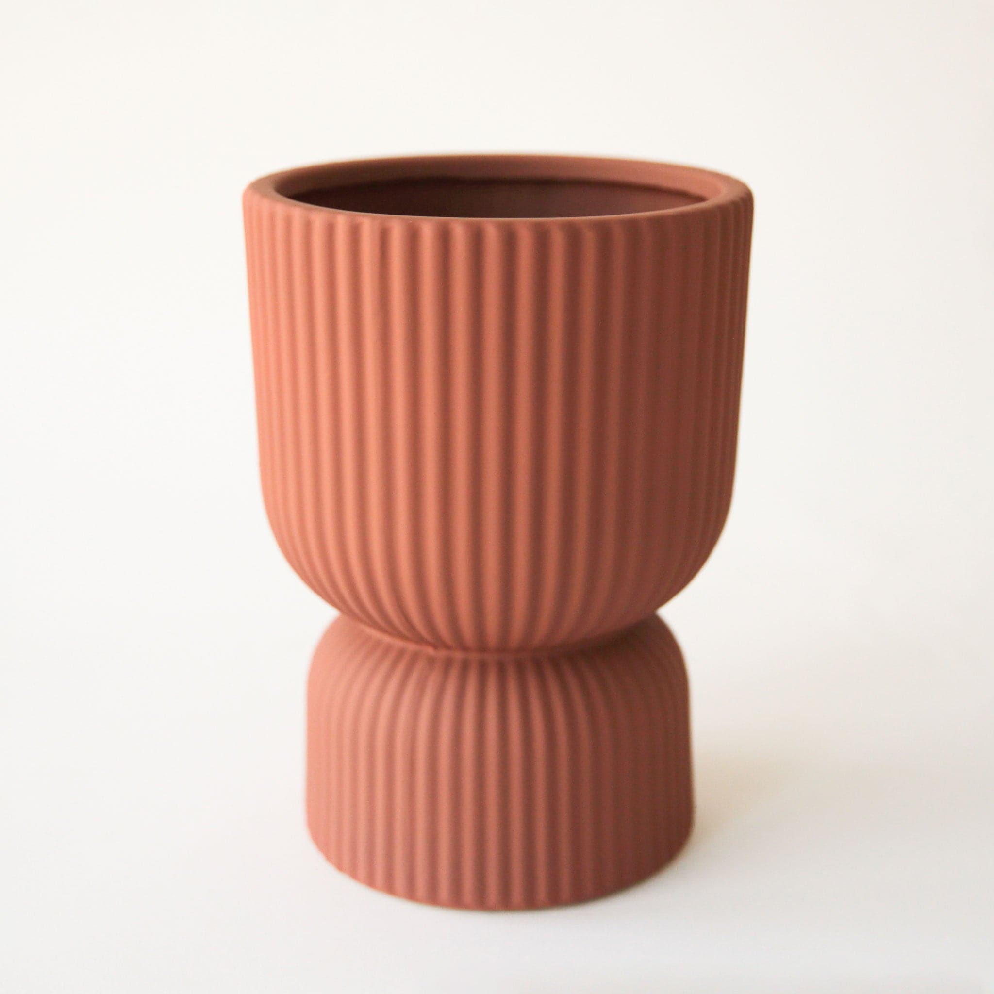On a white background is ribbed ceramic pedestal vase in a rust/brown shade. 