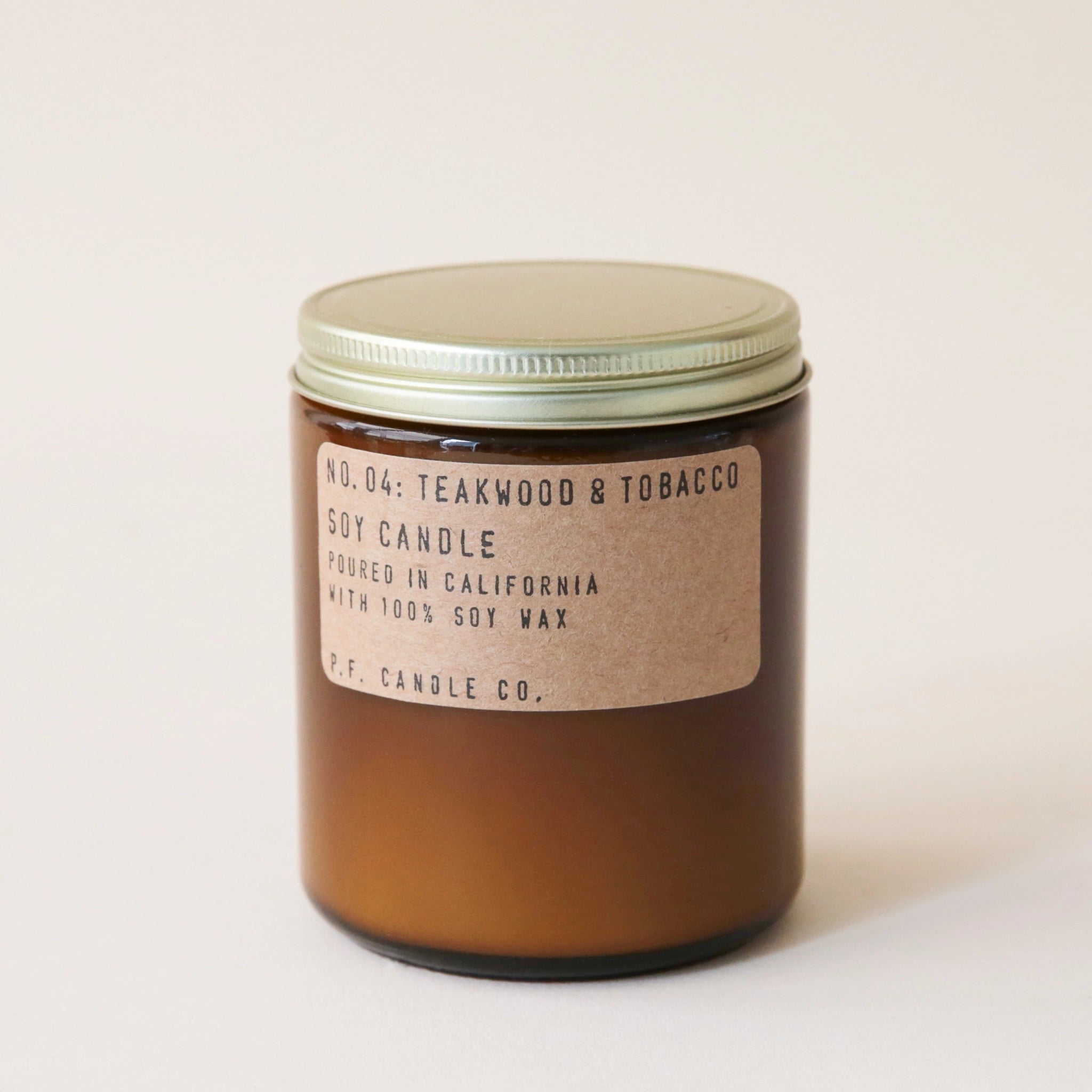 Against a white background is a dark amber, round jar. The jar has a gold lid. On the front of the jar is a brown, rounded, rectangular sticker with black text. The text reads ‘no.04 teakwood & tobacco. Soy candle. Handmade in California with 100% soy way. P.F. candle co.’