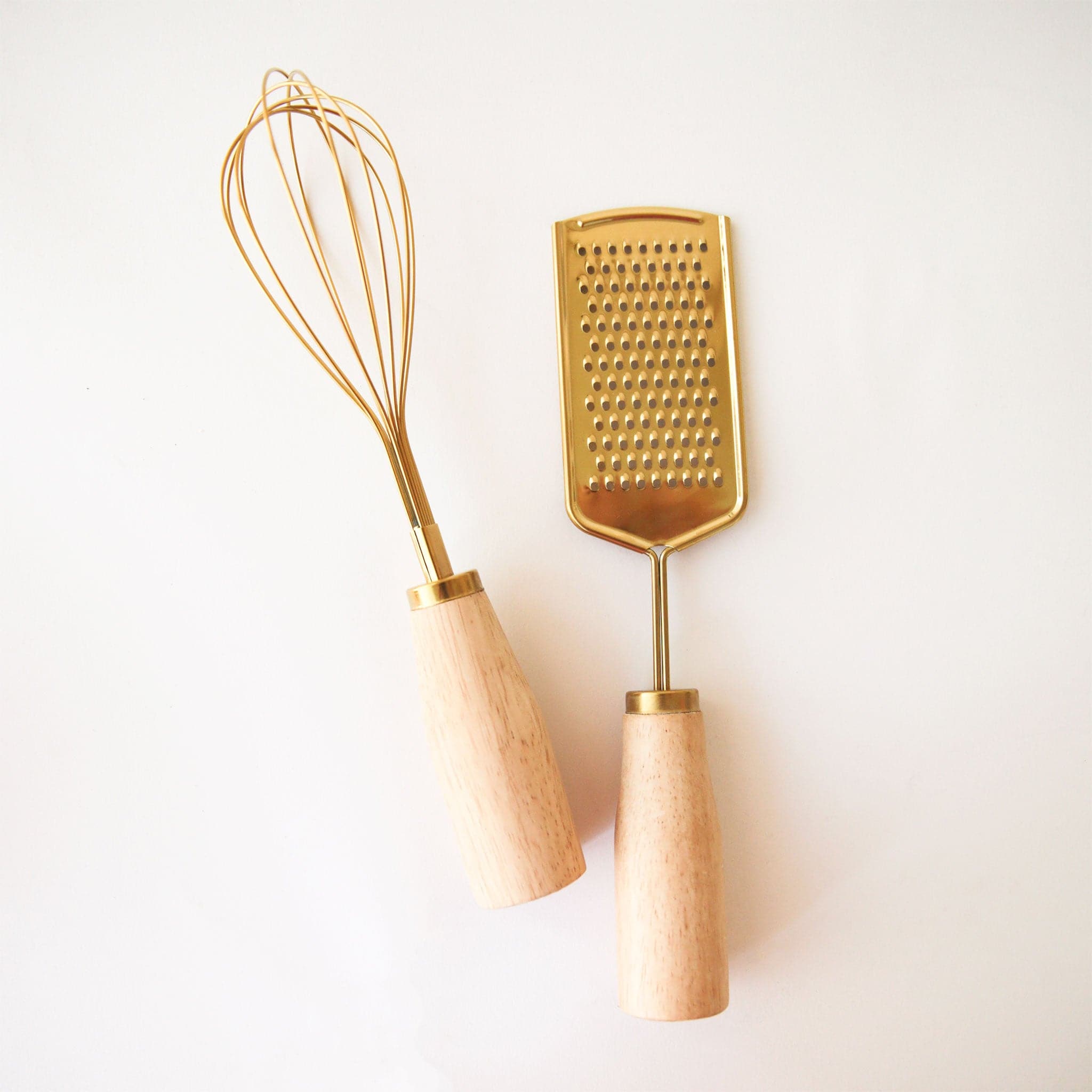On a white background is a gold whisk with a wood handle photographed with another kitchen tool. 