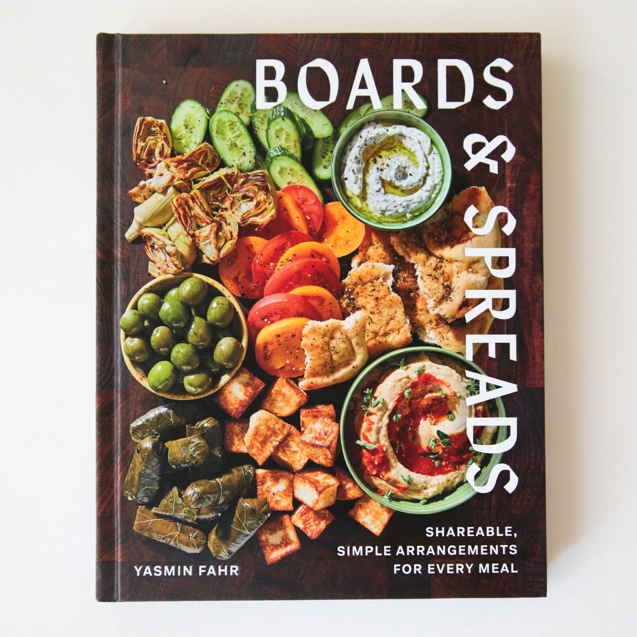 A book covering mimicking the look of a cutting board with a variety of charcuterie style foods, like olives, spreads, veggies and more. 