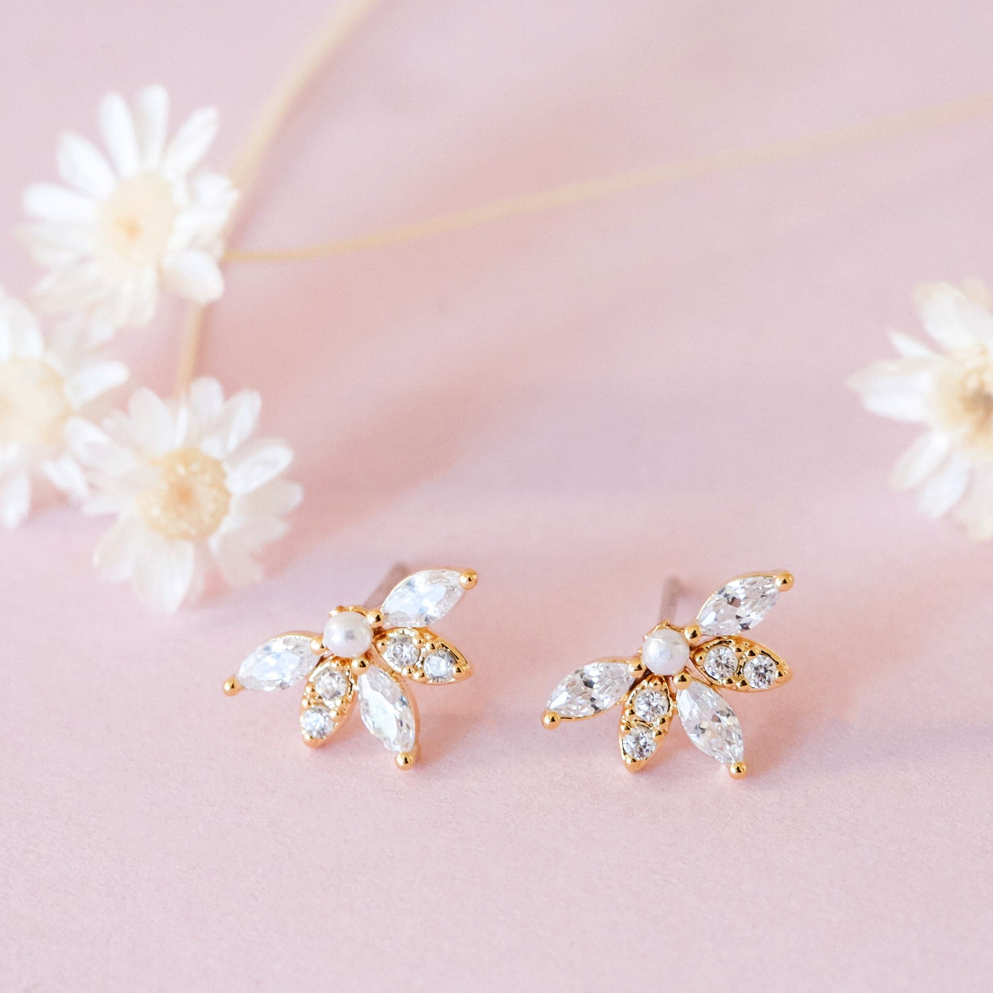 Gold metal stud earrings set with a pearl surrounded by a almond-shaped set crystal. Dried flowers are shown in the background.