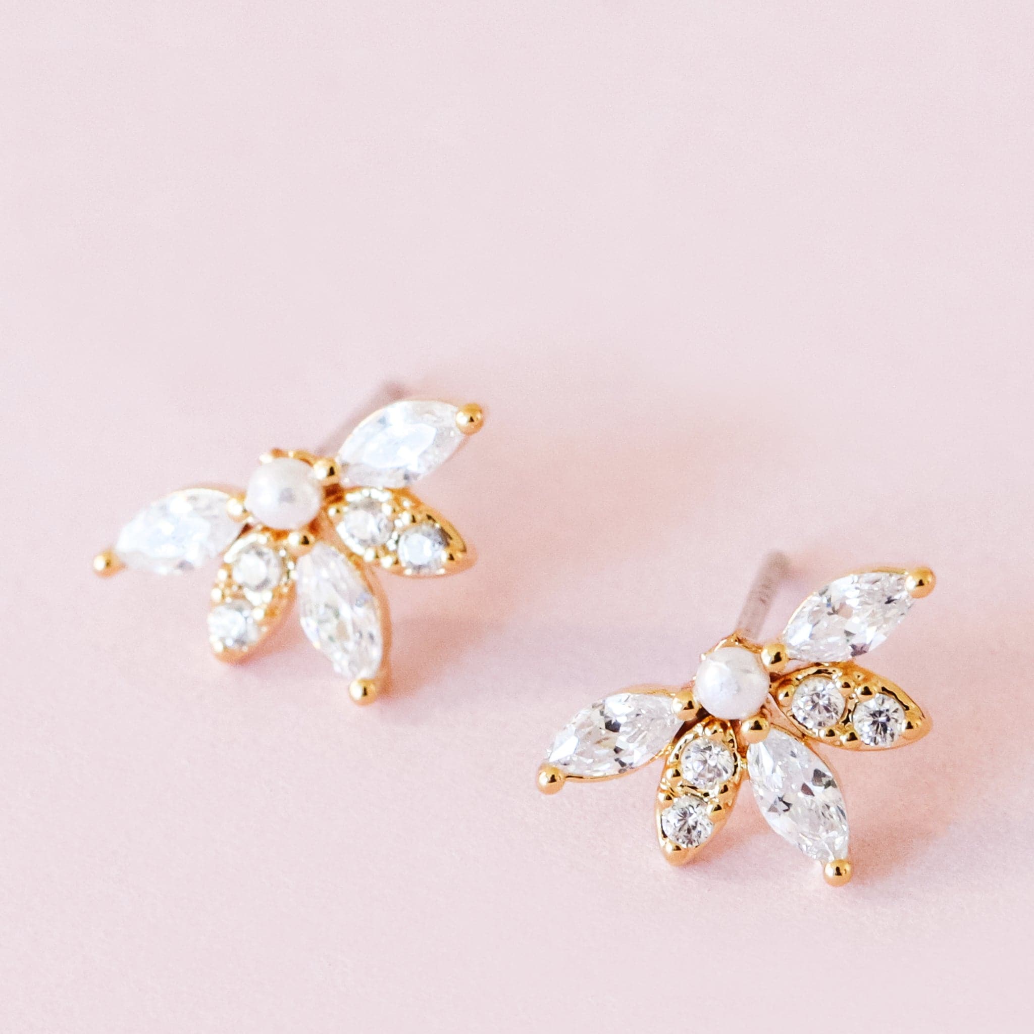 Gold metal stud earrings set with a pearl surrounded by a almond-shaped set crystal. Dried flowers are shown in the background.
