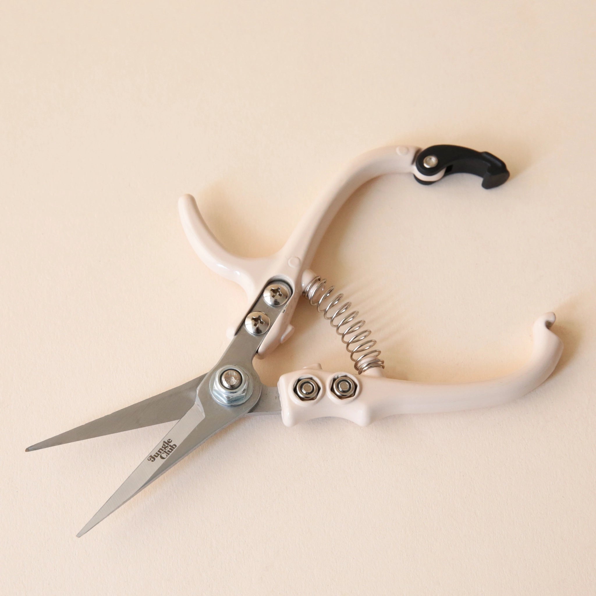 Pruning shears with a narrow tip, and light cream colored handle and a black clasp to connect when shears are not in use. There is small black text on the side of the shears that read, "Jungle Club".