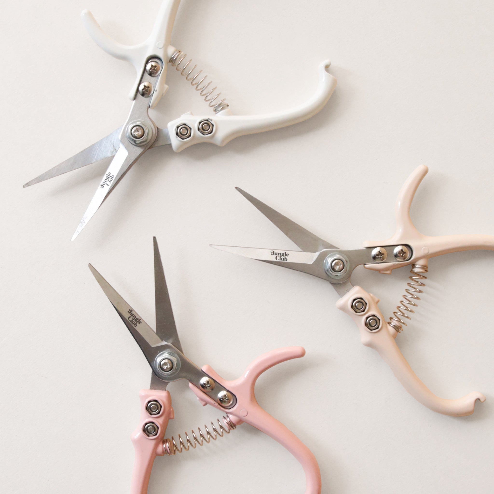 Pruning shears with a narrow tip, and light cream colored handle and a black clasp to connect when shears are not in use. There is small black text on the side of the shears that read, &quot;Jungle Club&quot; photographed here with the other colors available, pink and white.