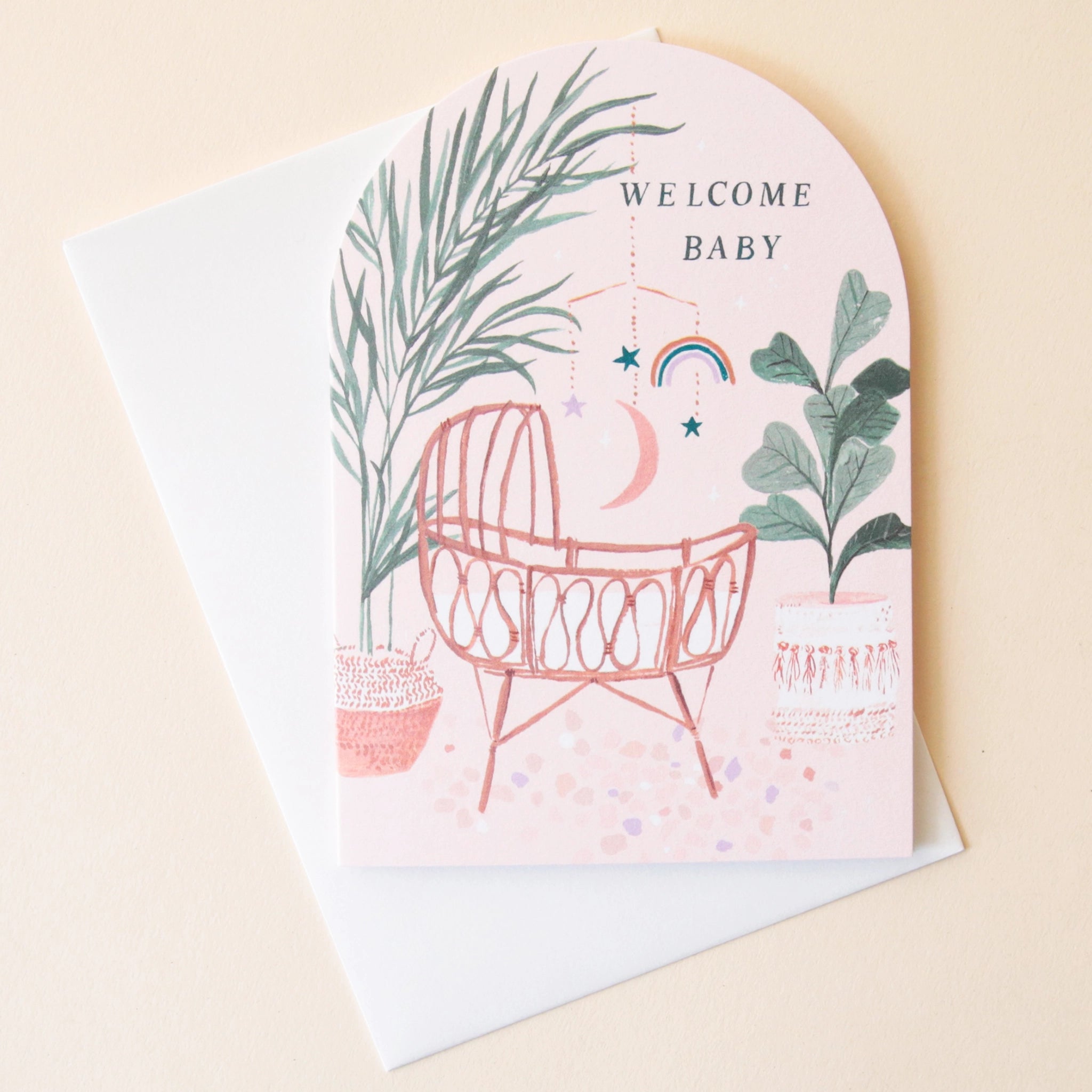 An arched greeting card with a sunset desert color pallet along with an illustration of a woven rattan baby&#39;s crib along with bohemian illustrations of lush natural foliage, bamboo, palm trees, and dried flowers and text at the top that reads, &quot;Welcome Baby&quot; in black letters.