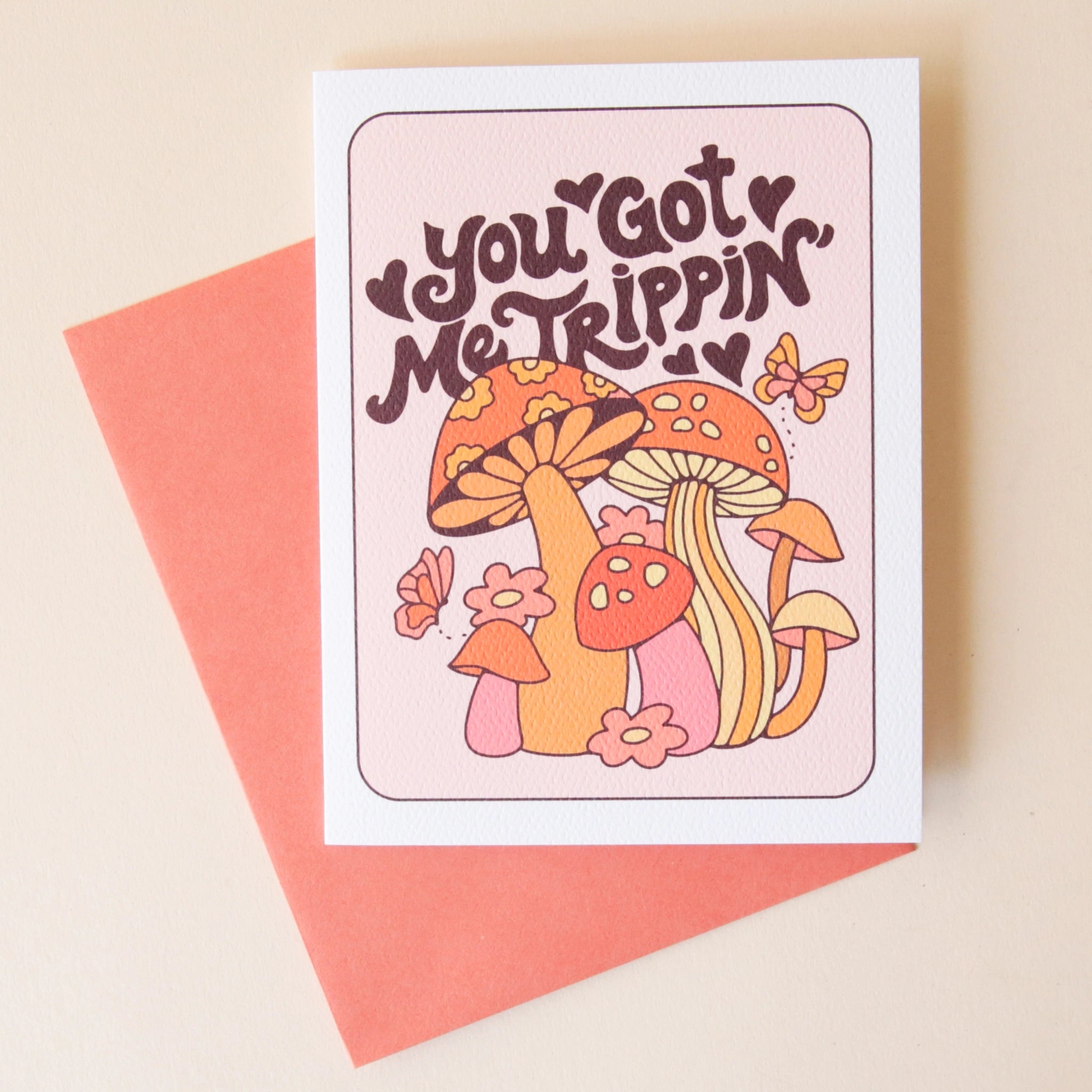 A white and light pink greeting card with mushroom and daisy graphics in the front along with groovy style font that reads, &quot;You Got Me Trippin&#39; in dark purple / brown letters and accompanied by a salmon pink envelope.
