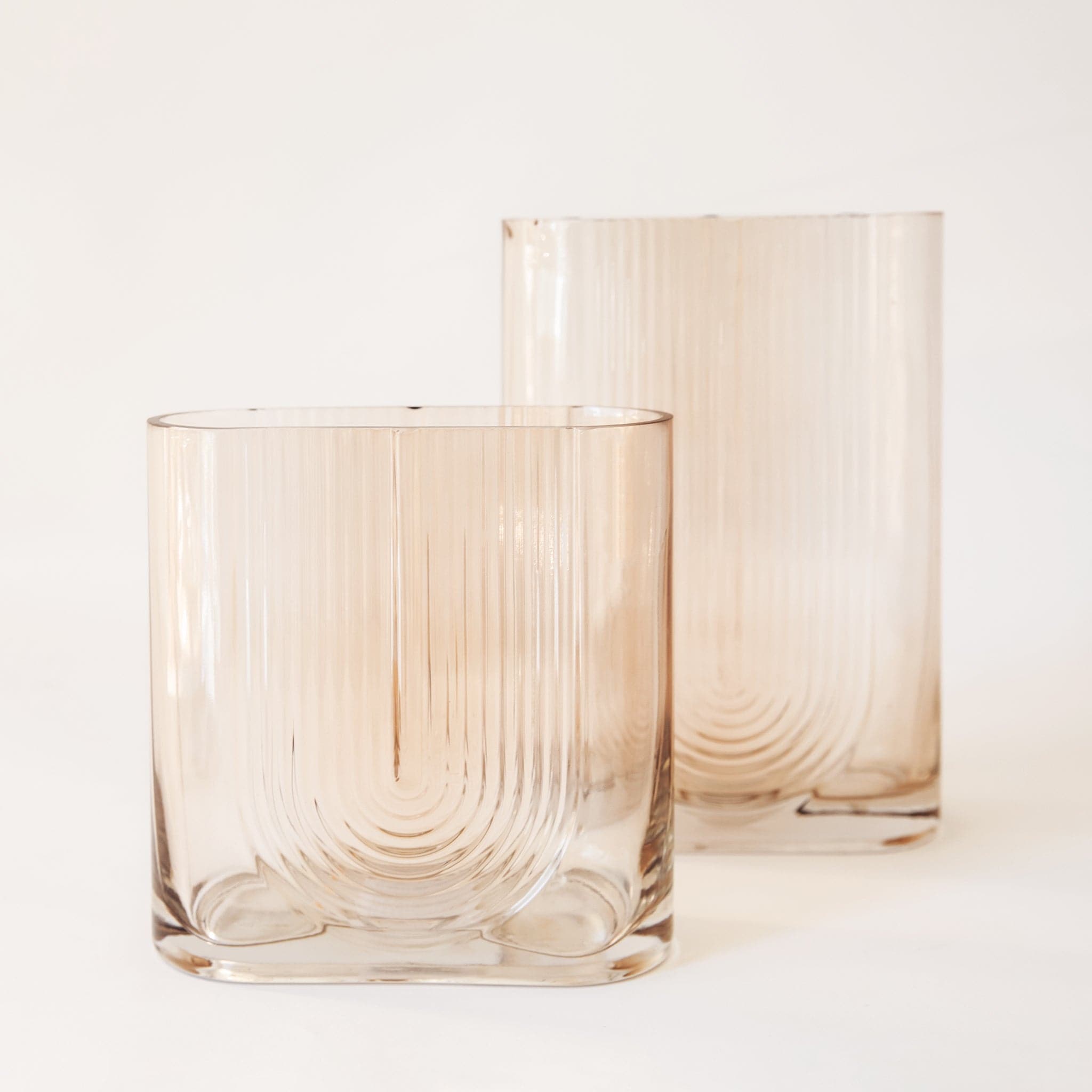 The morena vase in small and large alone. The morena vase has a unique shape. It is made from glass and has an oblong opening. The body of the vase is tall and angular resembling a square or rectangle, but with a pill-shaped opening the same width as the vase. There is a design on the front resembling a boho, upside-down rainbow design.