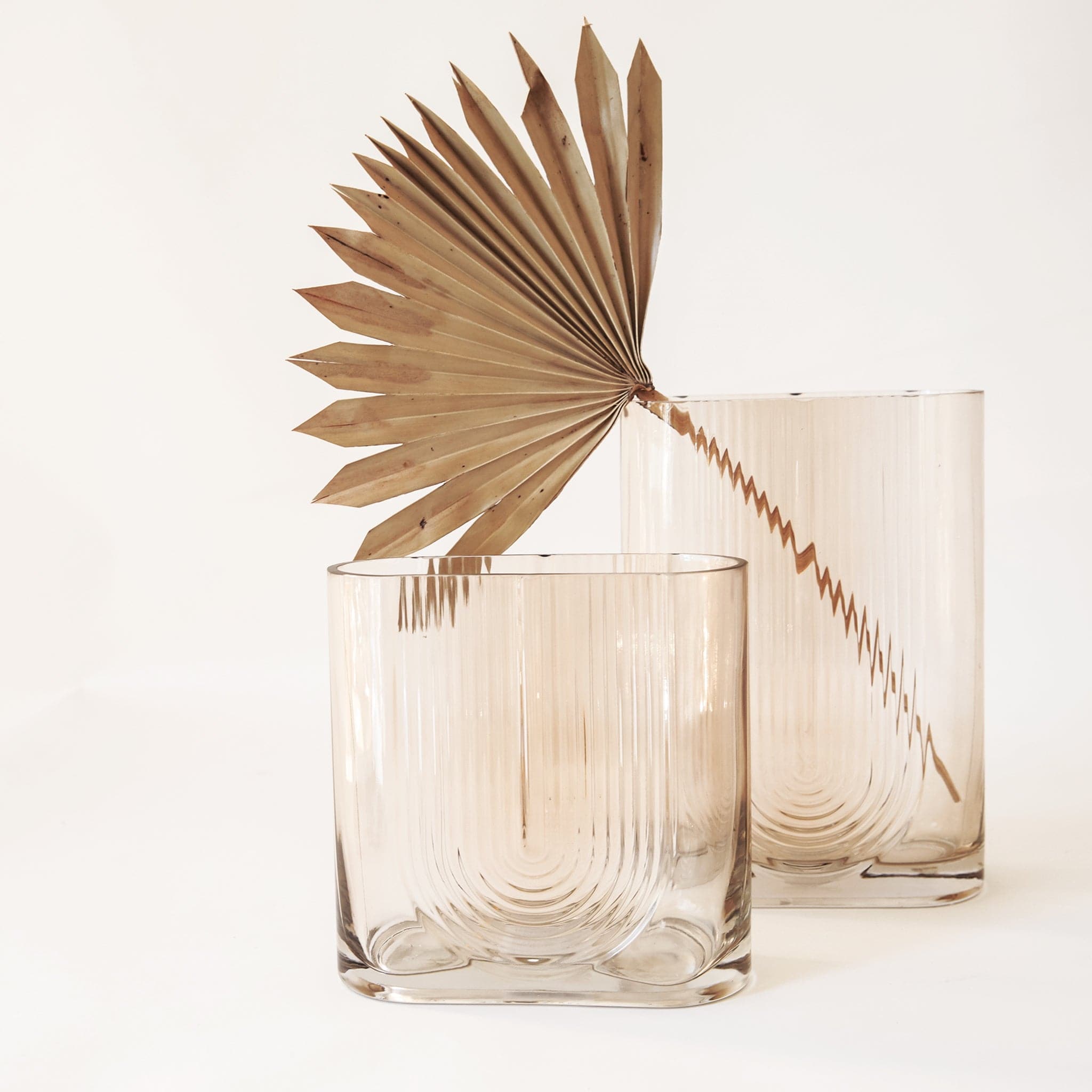 The morena vase in small and large with one dried palm fan in the large vase. The morena vase has a unique shape. It is made from glass and has an oblong opening. The body of the vase is tall and angular resembling a square or rectangle, but with a pill-shaped opening the same width as the vase. There is a design on the front resembling a boho, upside-down rainbow design.