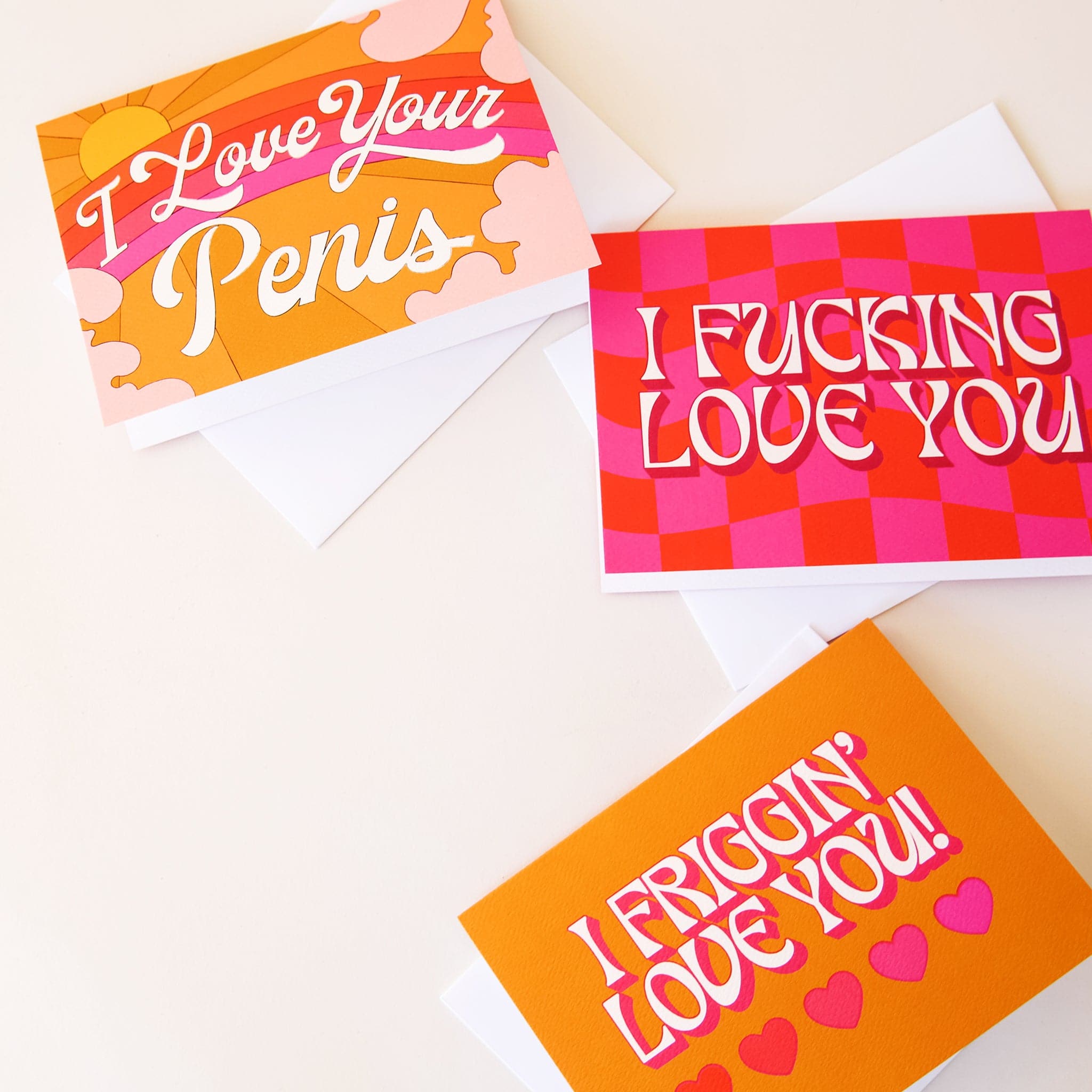 Three of our Sunshine Studios cards on a white background. Each card has bright, rich color, reminiscent of the 70's with warm tones. The first card says "I love your penis". It features a rainbow, sun, and clouds. The second card has a hot pink and deep orange wavy checker background. The script on the card is white and reads "I fucking love you". The third card has a solid gold color and reads "I friggin' love you!". There are five little hearts, each a different color.