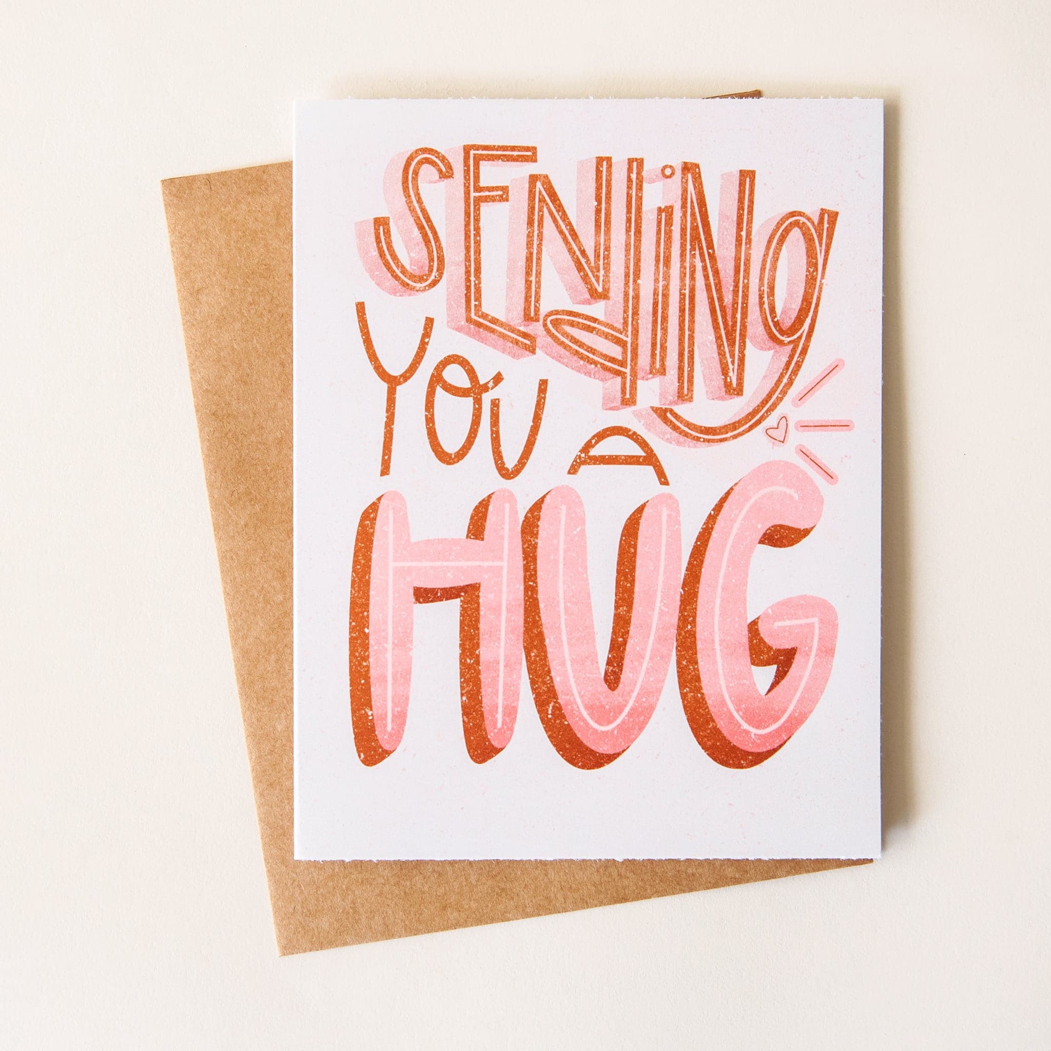 An adorable card with fun, playful font reading, &quot;Sending you a hug&quot;. The font takes up the whole card, and has rust and light pink coloring. There is a designed texture on the font, giving it a hand screened look.