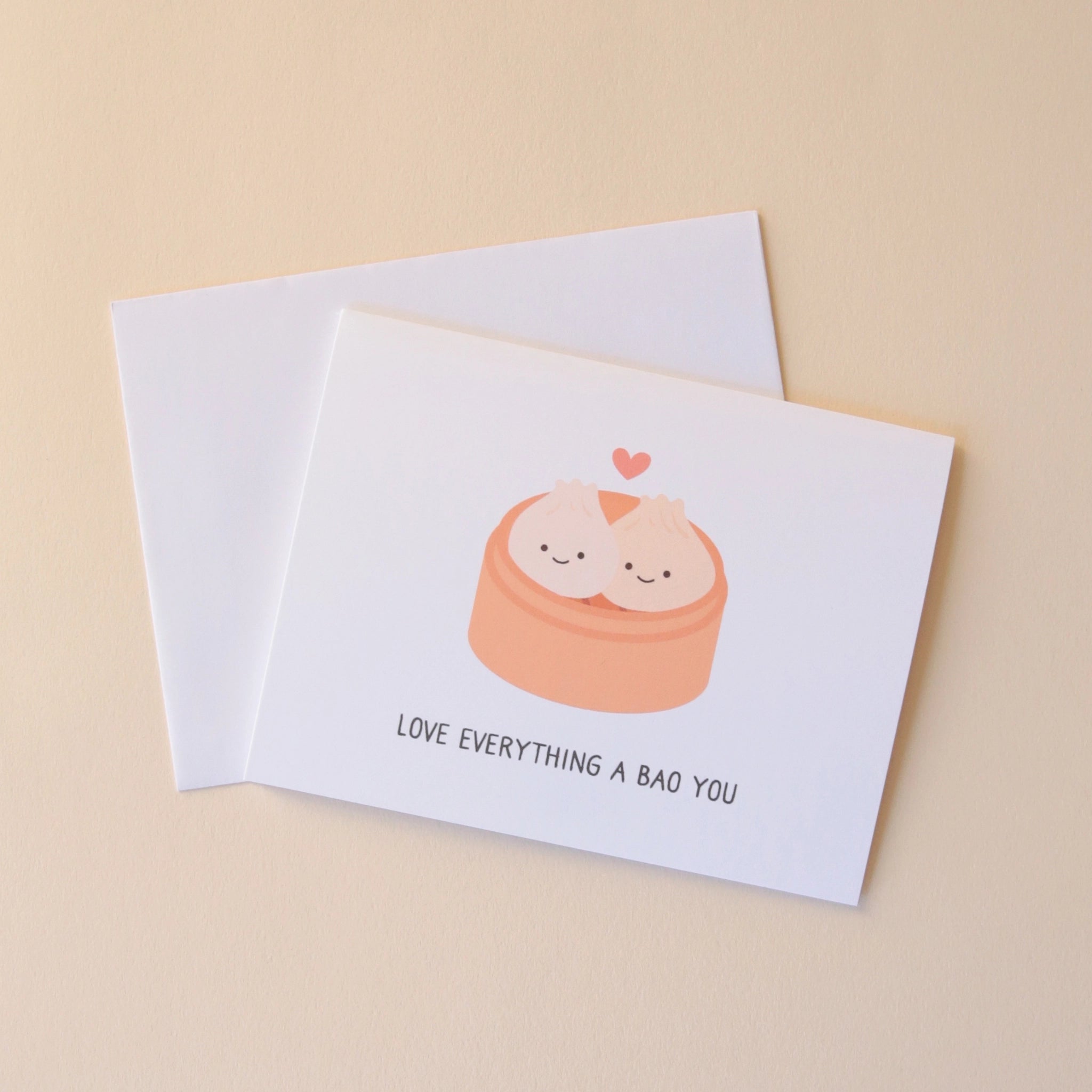 A white card with two bao buns with smiling faces in a steamer with a small pink heart above them and text underneath that reads, "Love Everything A Bao You" in black letters.