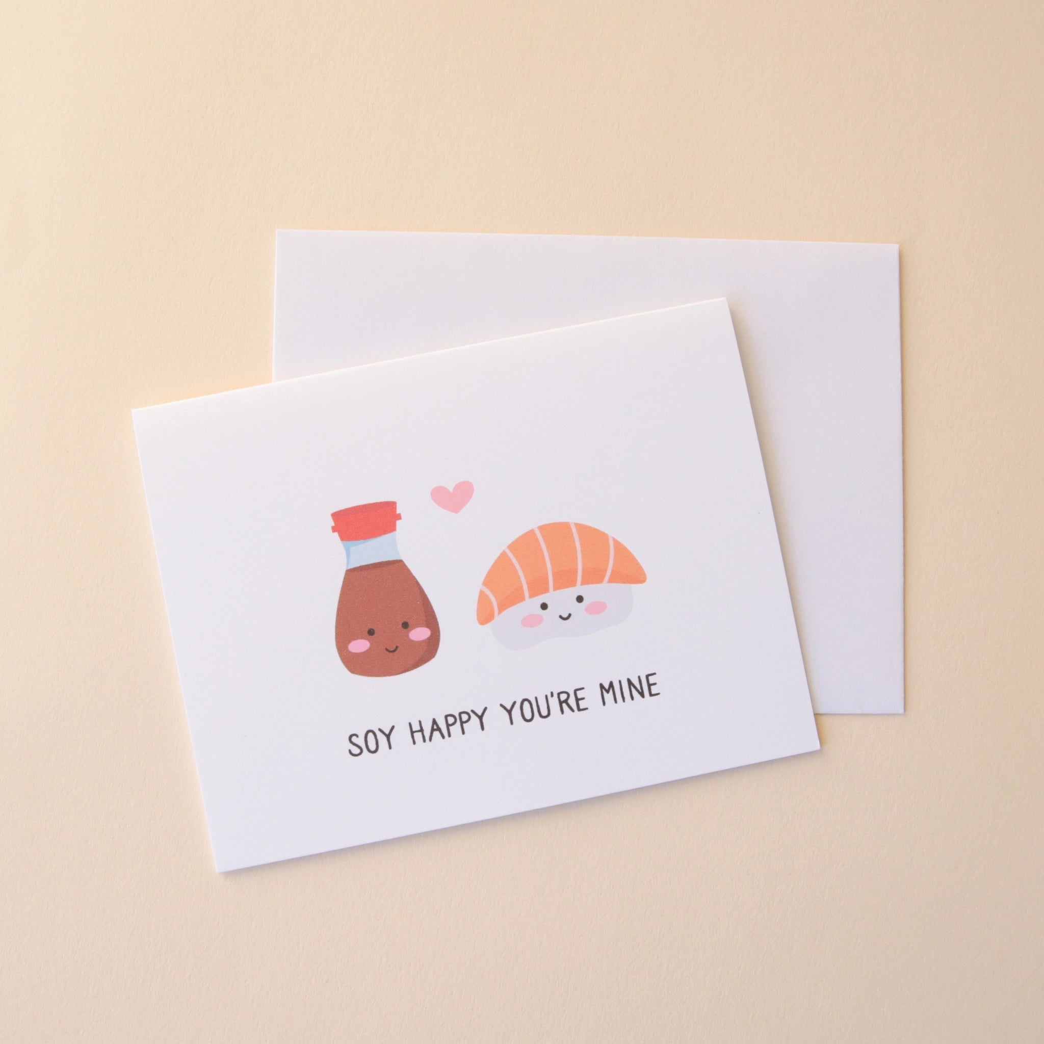 A white card with an illustration of a salmon sashimi and soy sauce both with smiling faces and a small pink heart in between them as well as text underneath that reads, "Soy Happy You're Mine" in black letters.