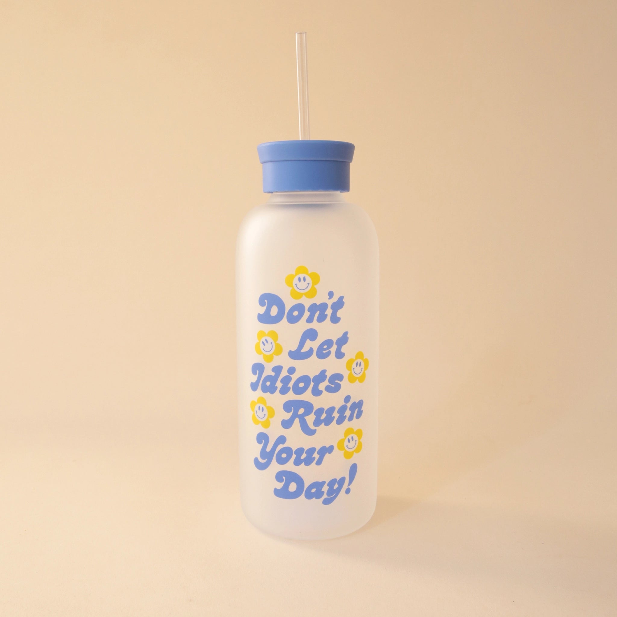 On a tan background is a clear water bottle with a blue lid and blue text that reads, &quot;Don&#39;t Let Idiots Ruin Your Day&quot; with smiley daisies all around it. 