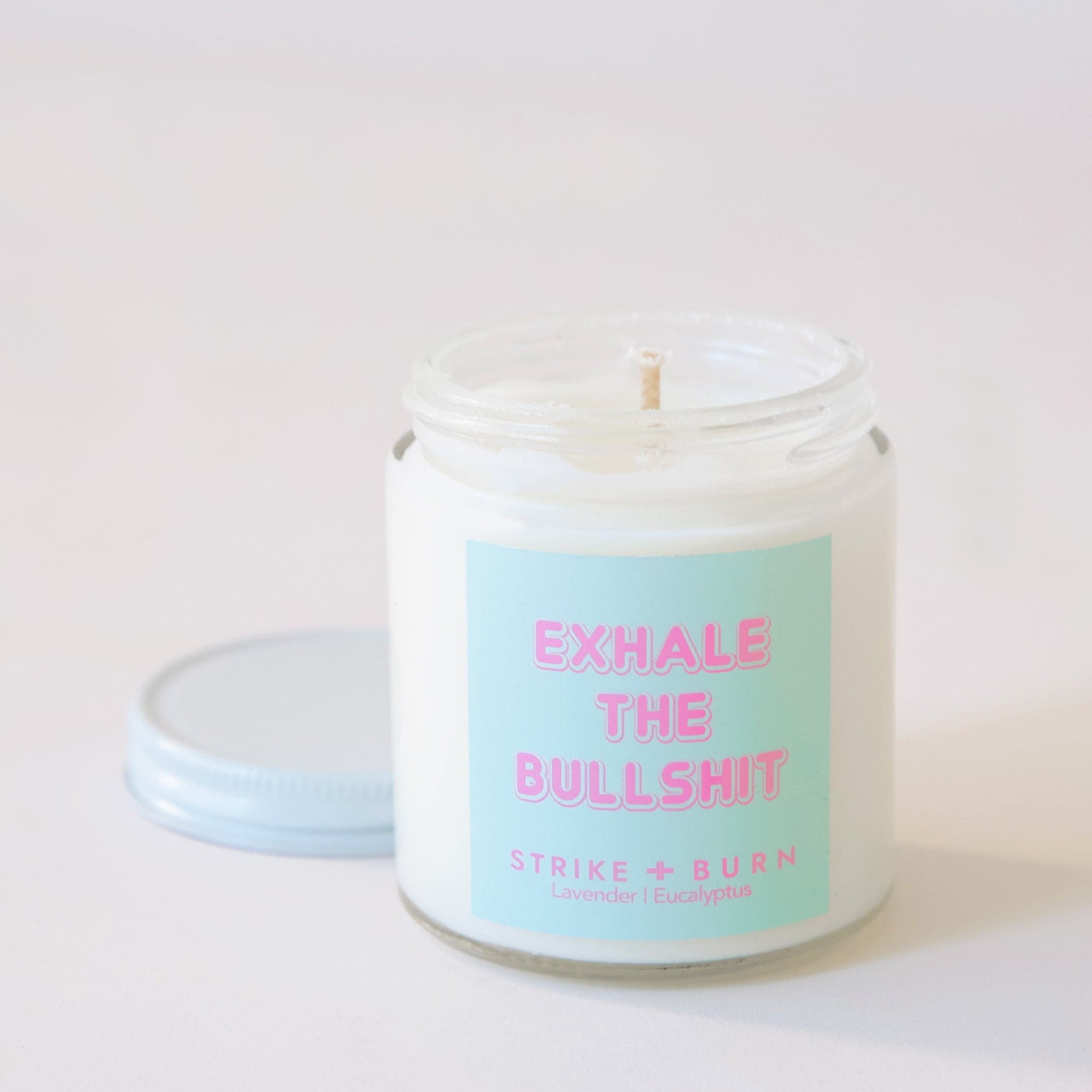 Clear, glass jar candle with white metal lid. Label says "Exhale the bullshit" in pink with a aqua colored background.