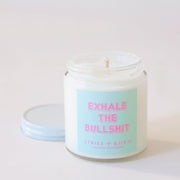 Clear, glass jar candle with white metal lid. Label says "Exhale the bullshit" in pink with a aqua colored background.