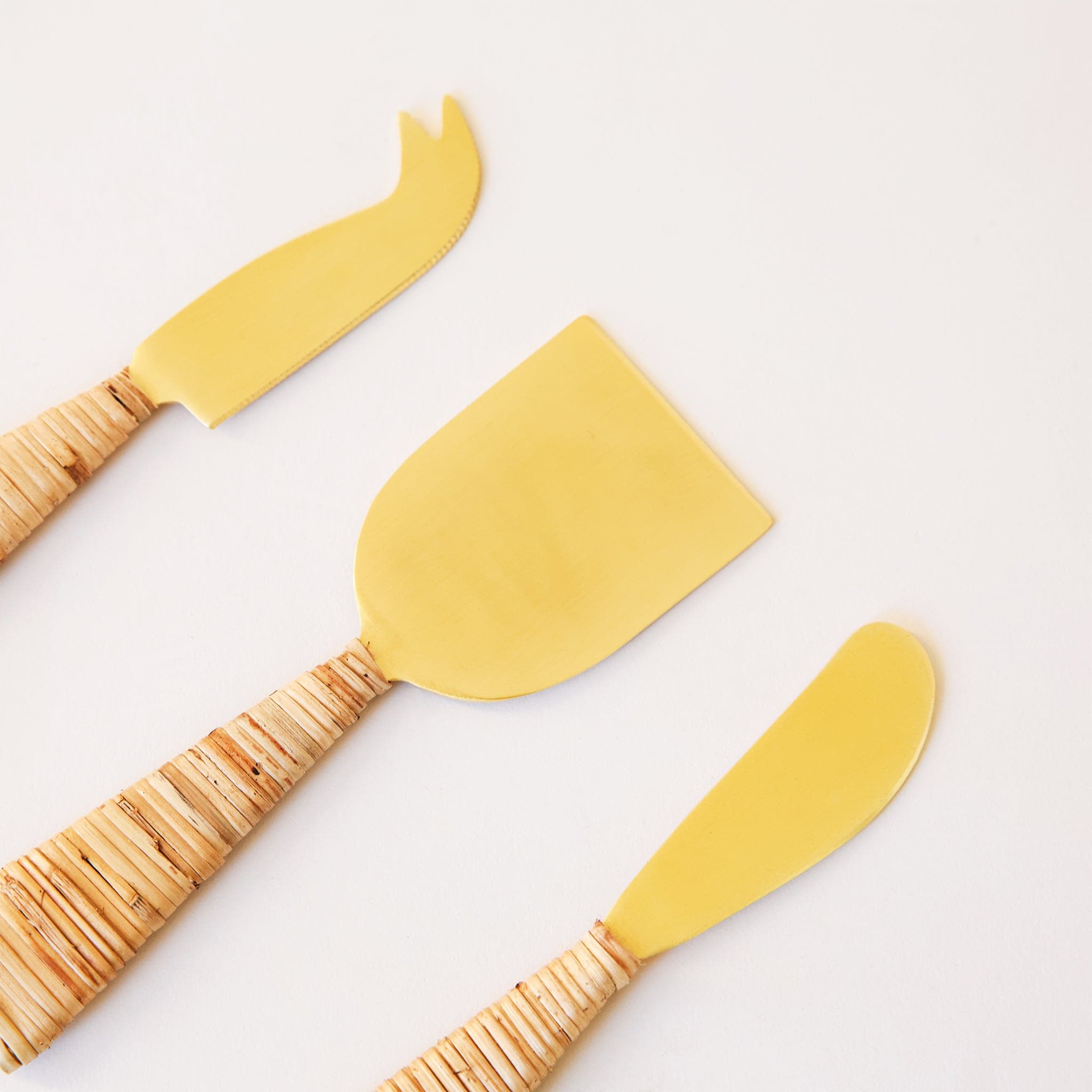 Gold cheese knives wrapped with natural rattan handles. A hatched design is on one side of the knives, while the opposite side is horizontally wrapped. 
