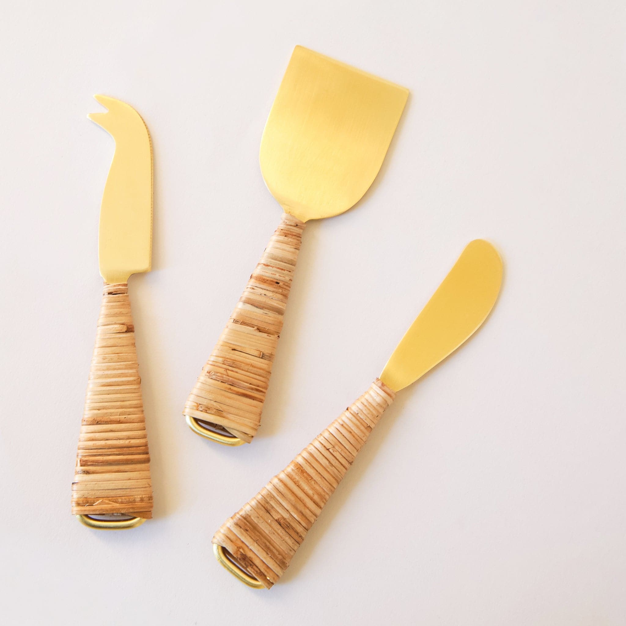 Gold cheese knives wrapped with natural rattan handles. A hatched design is on one side of the knives, while the opposite side is horizontally wrapped. 