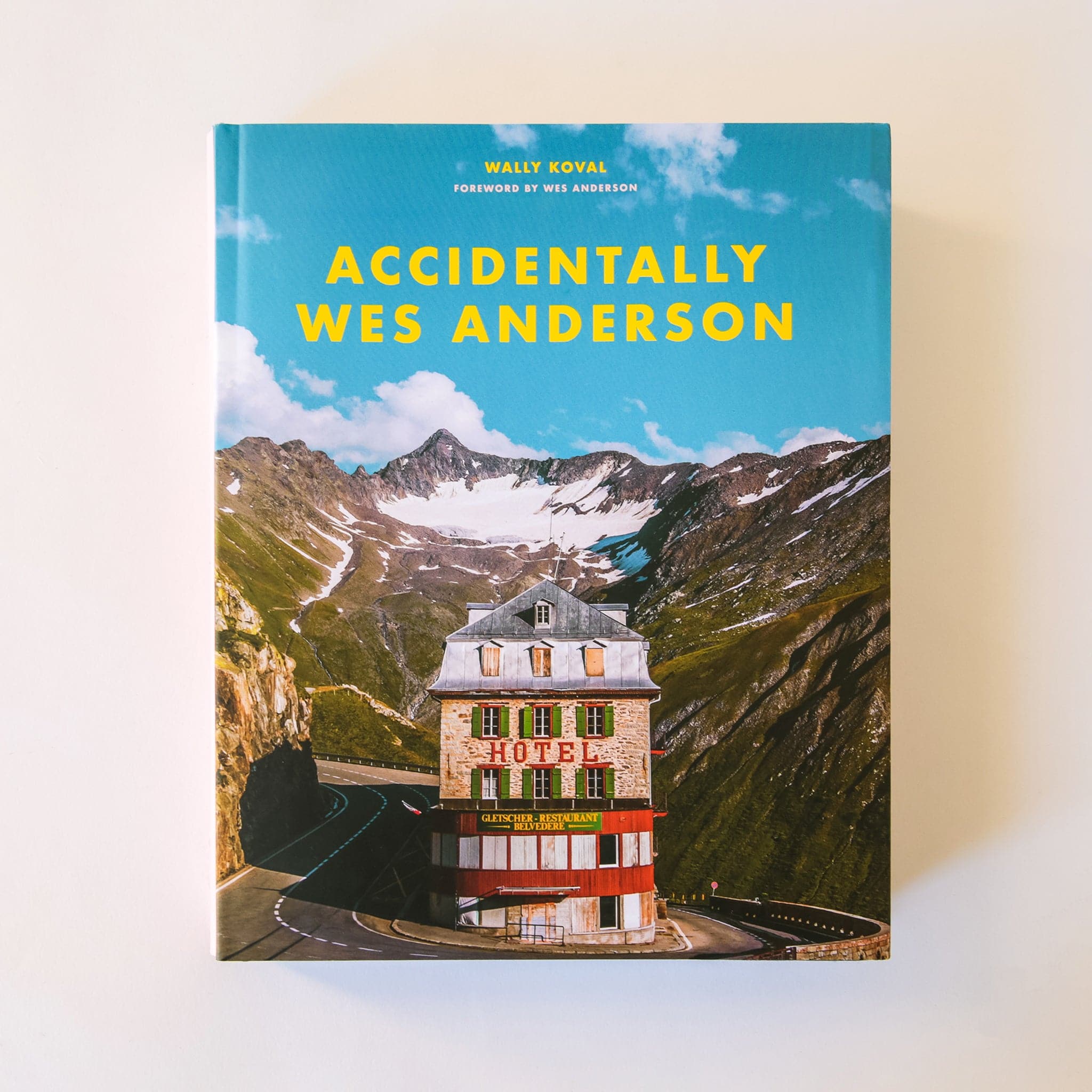 The cover of Accidentally Wes Anderson. It features an iconic scene with the Swiss Alps and a curving road around a brick hotel. It uses the color palette Wes Anderson is known for with the use of vivid warm, bright colors.