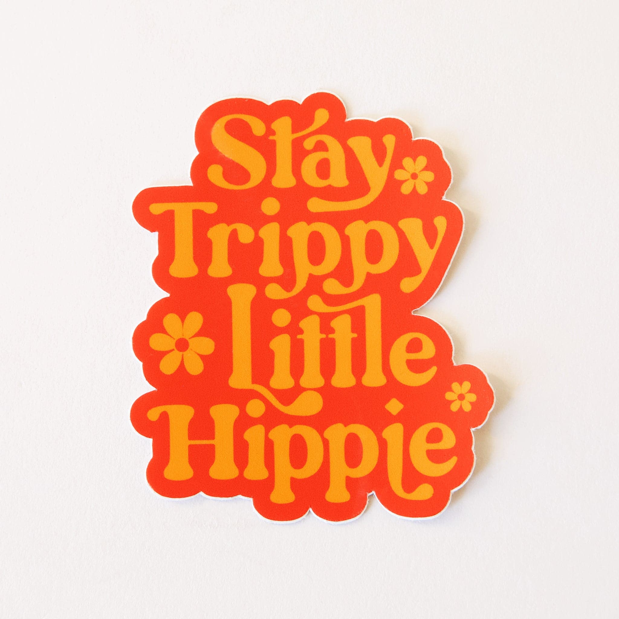 In front of a white background is a bright orange sticker with light orange text. The text reads ‘stay trippy little hippie.’ The words are stacked one on top of another. There are three orange flowers; one by the word ‘stay,’ one by the word ‘little’ and the last is by the word ‘hippie.’