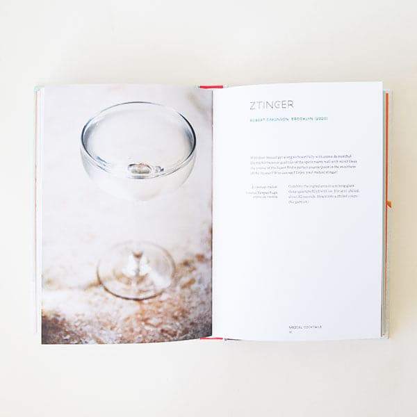 On a neutral background is the book opened to a page with a photo of a cocktail on the left and a description and recipe on the right. 