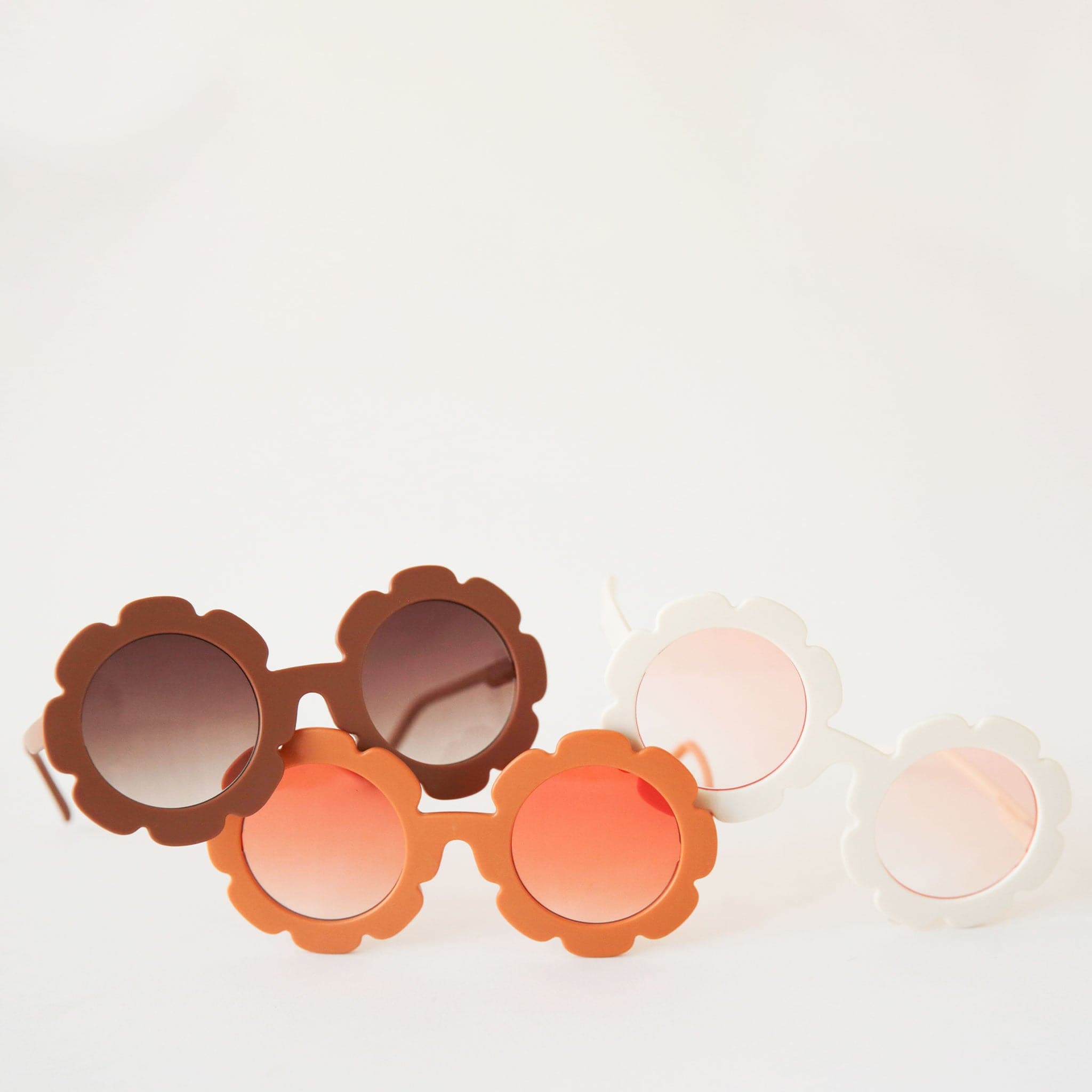 Brown flower shaped sunglasses with light brown circle lenses in the center photographed here with the other color ways available. One is an orange frame with a light orange lens and the other a white frame with a light pink lens.