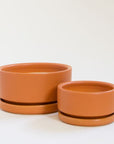 Two terracotta orange low bowl planting pots, both complete with water trays. Both sit empty besides each other.