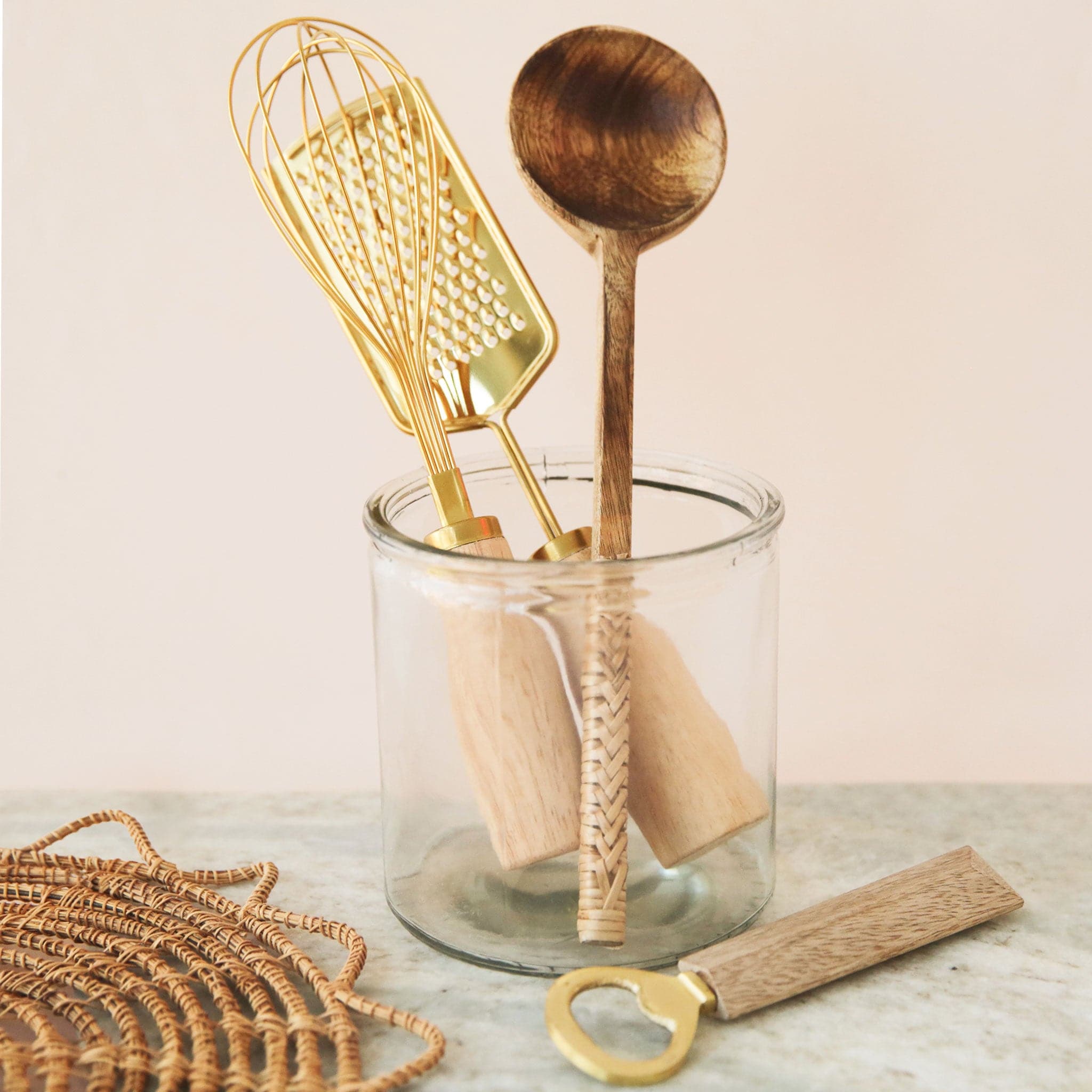 On a white background is a gold whisk with a wood handle photographed with other kitchen tools. 